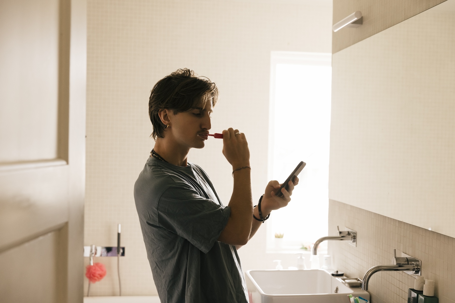 Side view of young man checking his phone while brushing teeth in bathroom at home