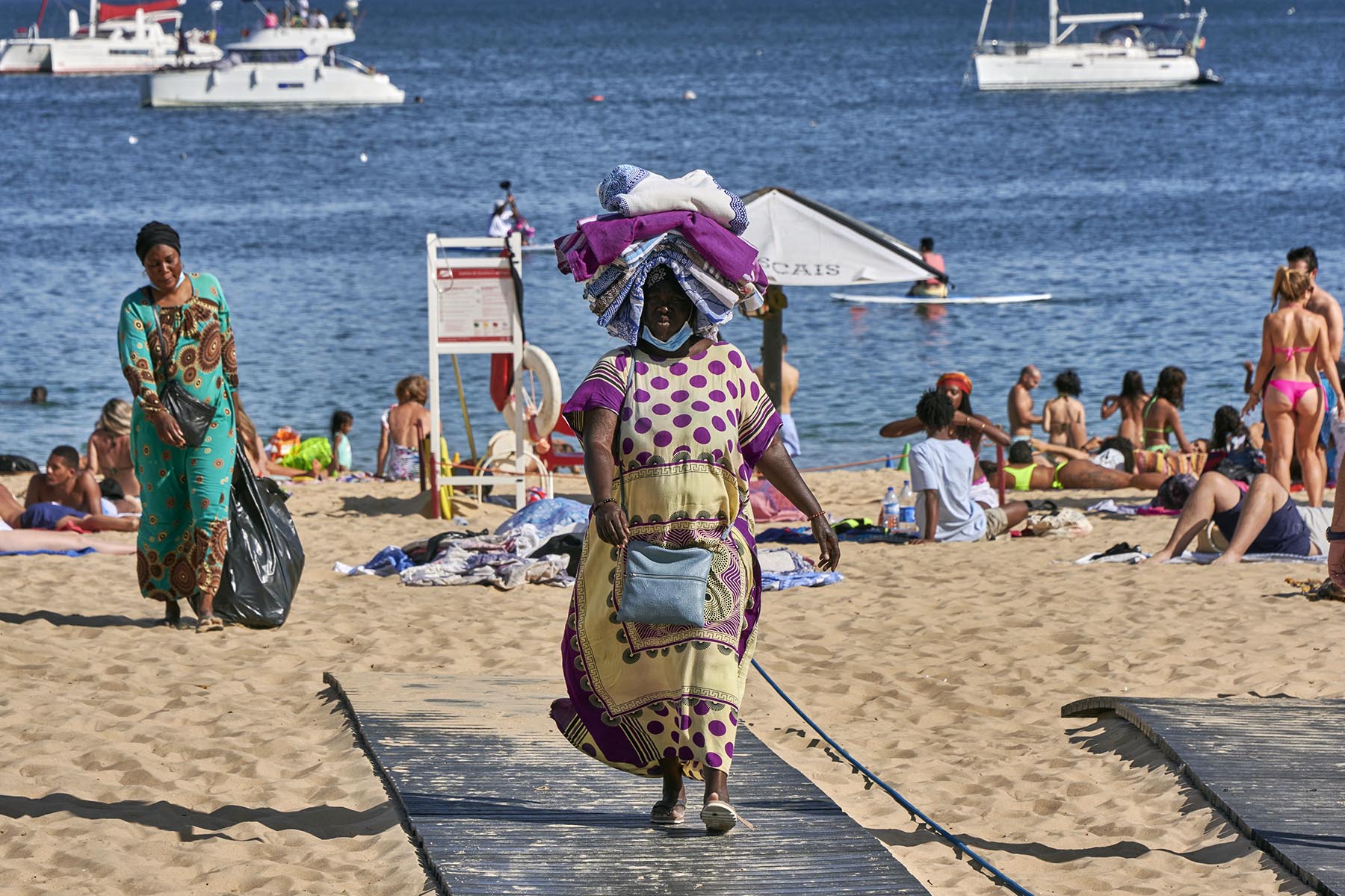 African vendors is carrying beach towels on his head to sell to beachgoers on a sunny day at Praia da Duquesa in Cascais, Portugal.