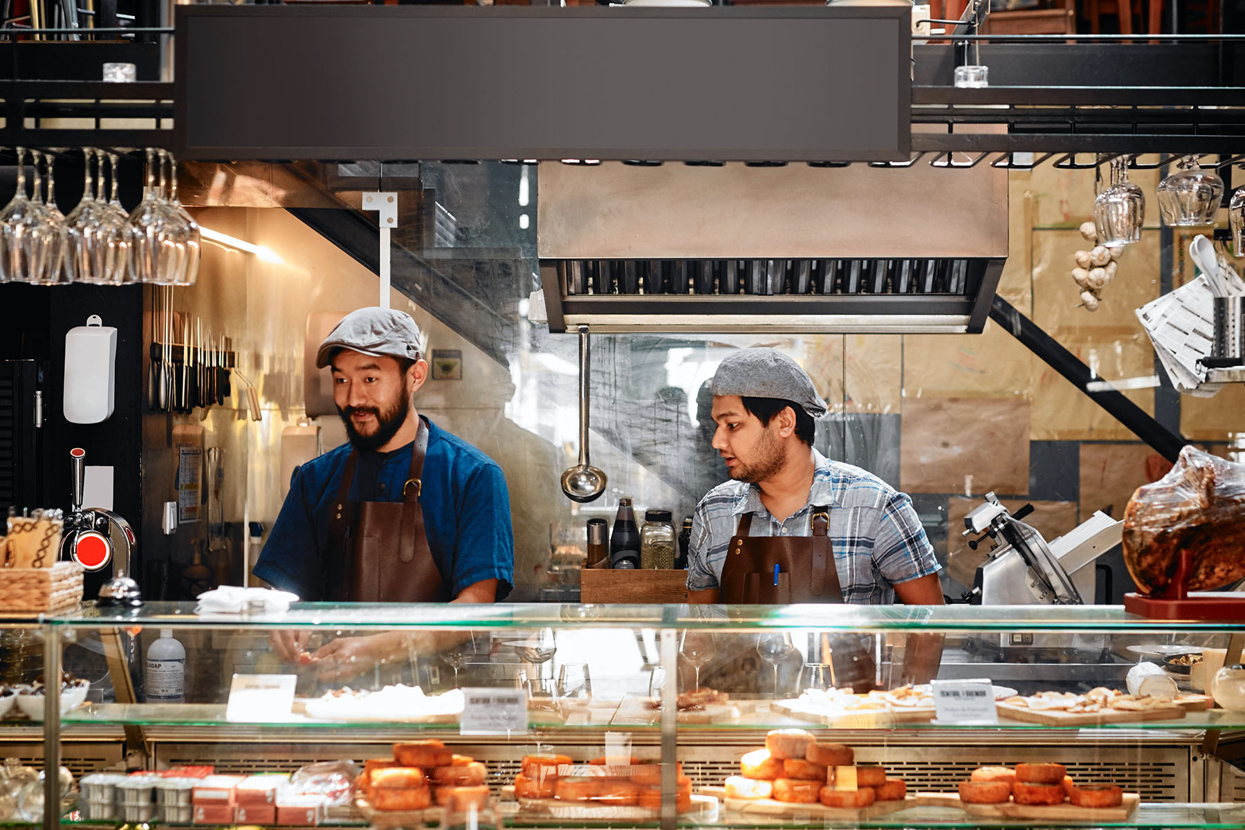 Two men working behind the counter of a food store or cafe. One is caught staring while the other one is looking at what he's making.