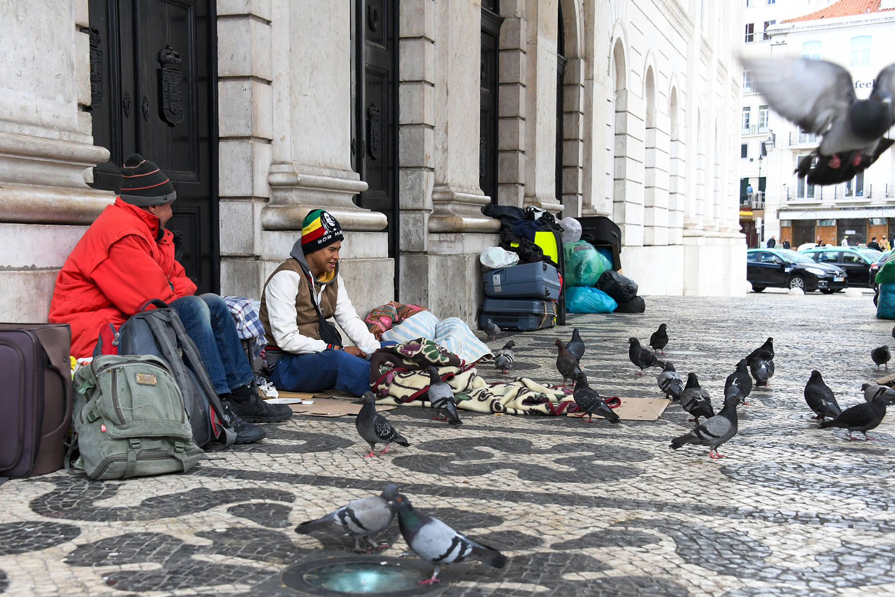 Two houseless people sitting on the ground at the National Theatre in Lisbon. All their possessions are piled around them. They're smiling as they are surrounded by pigeons.