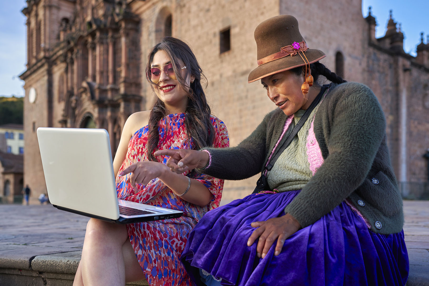 Two women are sitting at a town square, looking and pointing at a shared laptop. One is dressed in modern clothes, the other wears traditional Quechua (indigenous to Peru) dress.