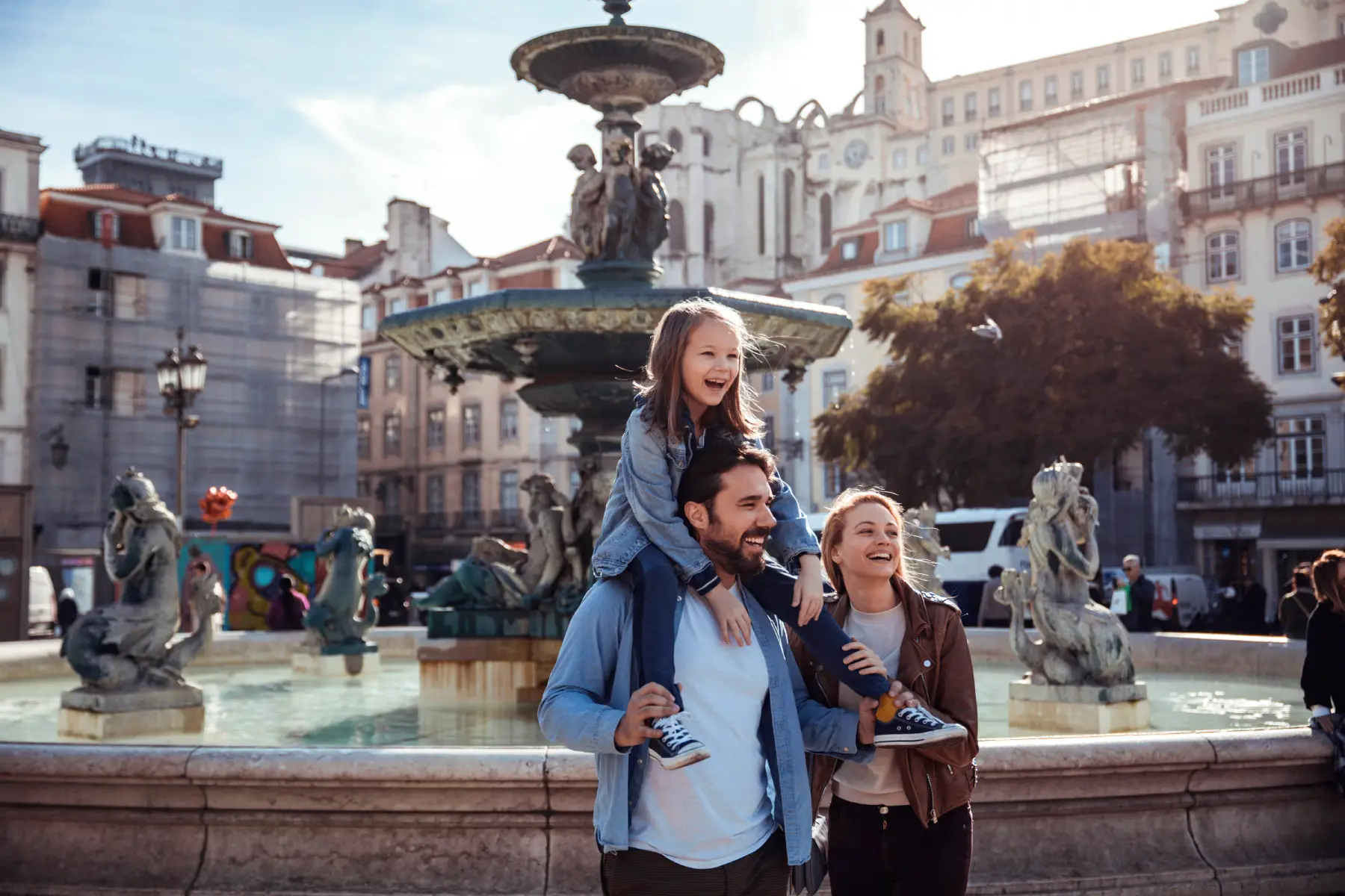 a little girl sitting on her father's shoulders as he walks past a water fountain in Rossio Square in Lisbon, with the mother walking alongside them