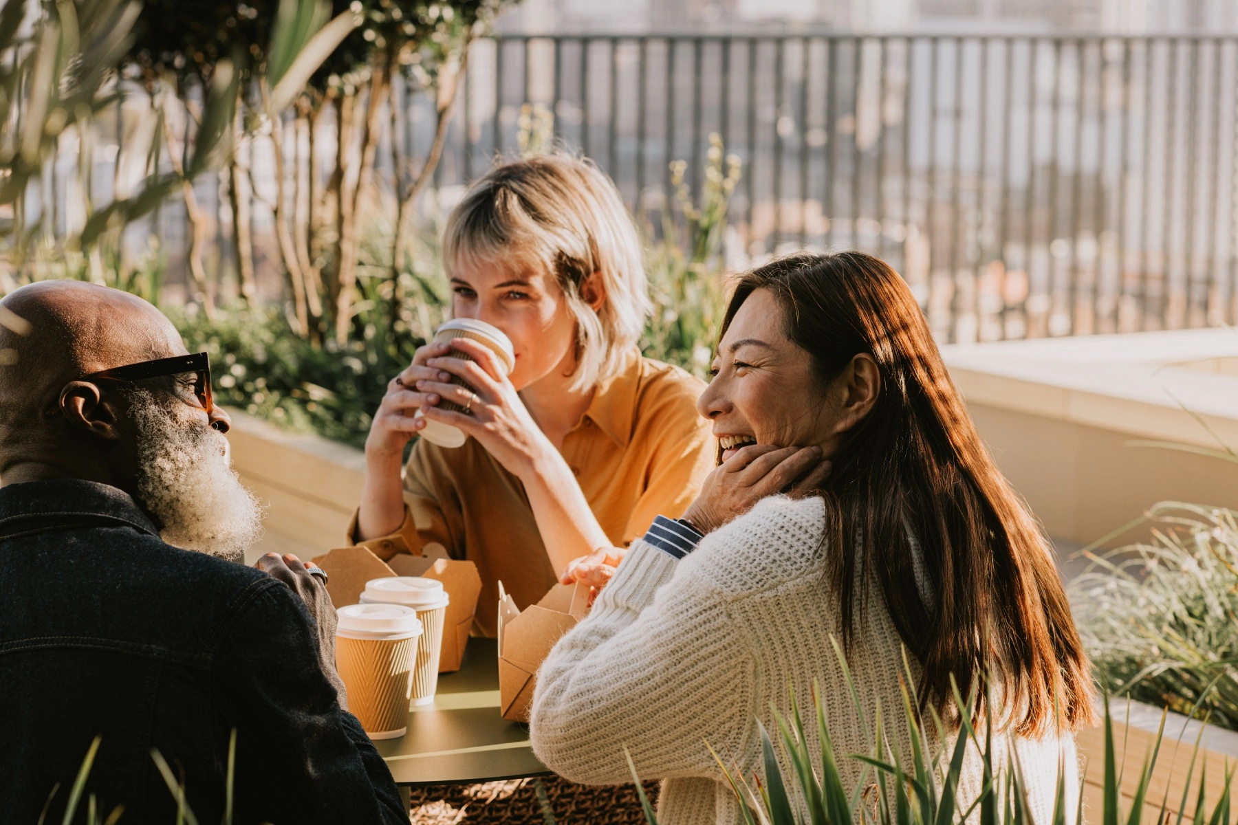 a group of three friends sitting outside in the sunshine, drinking coffee and chatting
