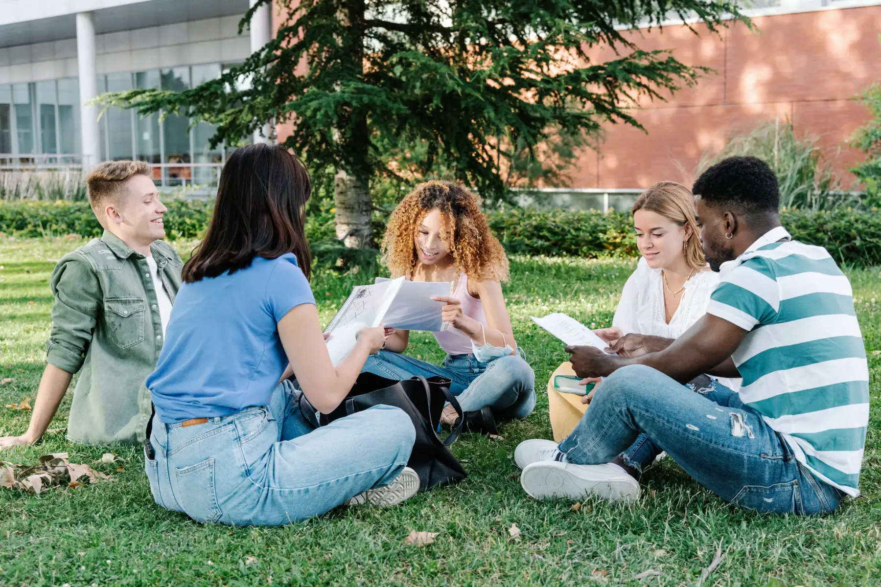 a group of young diverse men and women in casual clothes reading papers while sitting on lawn outside university building and doing homework together