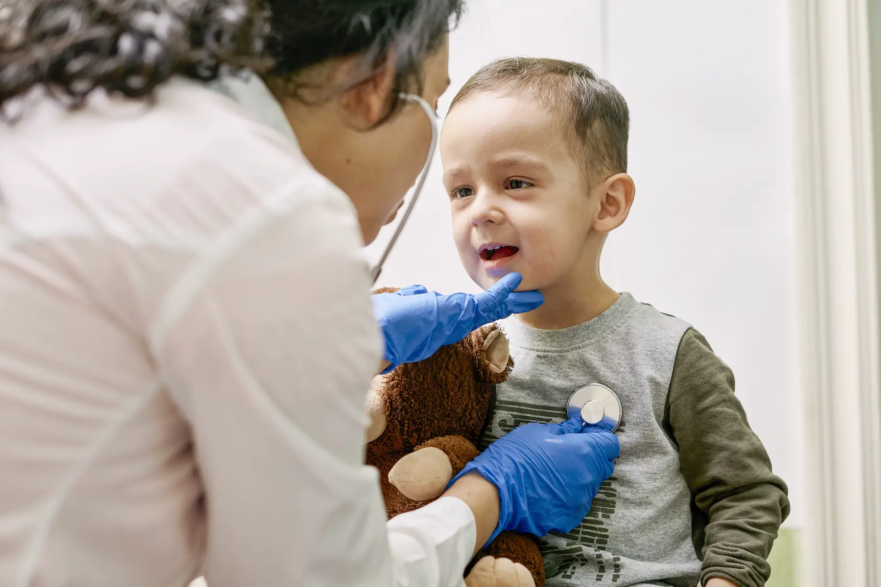 A little boy holds a teddy bear and opens his mouth at the pediatrician who is listening to his heart with a stethoscope.