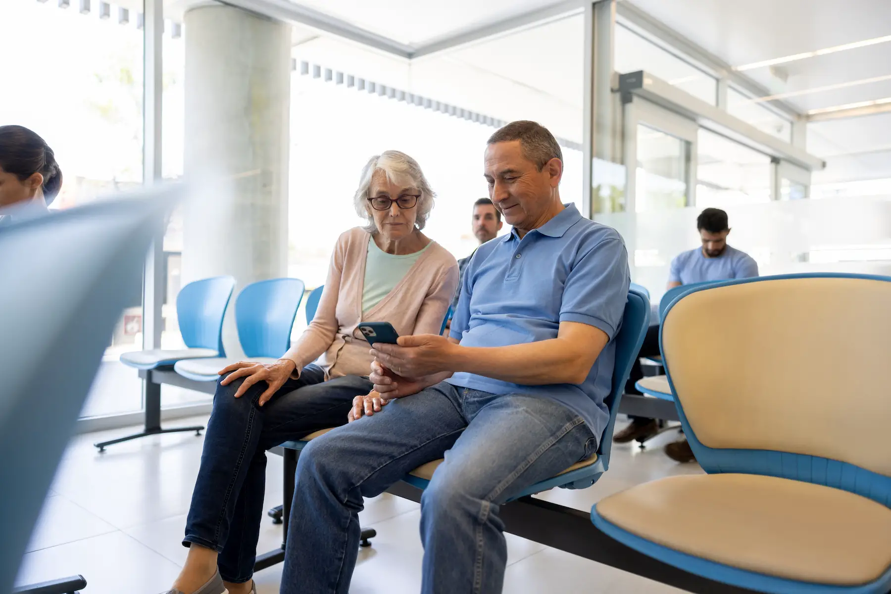 An older couple sits together in the waiting room at the hospital while looking at something on his mobile phone