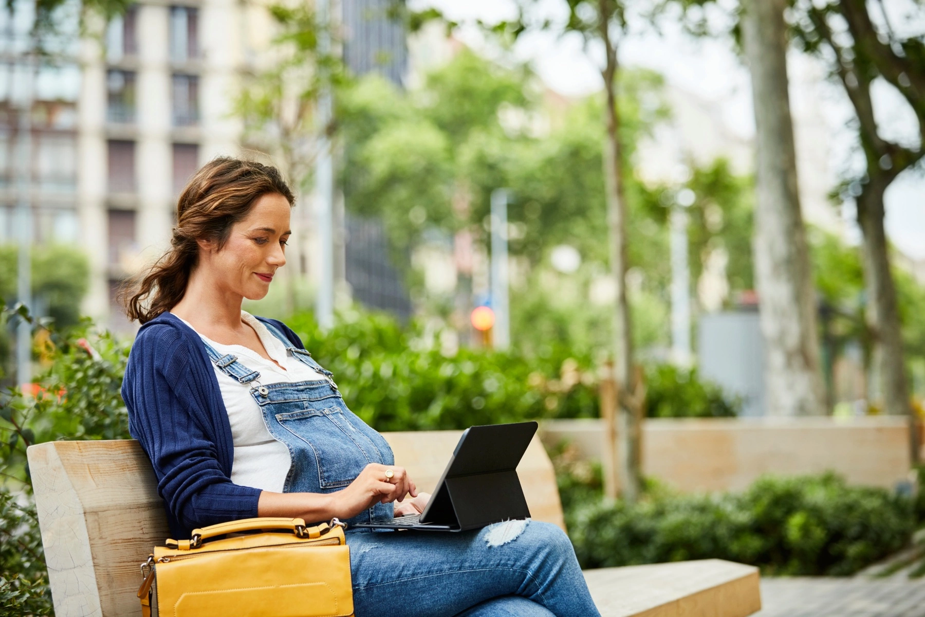 a pregnant woman using a laptop while sitting on bench in a park