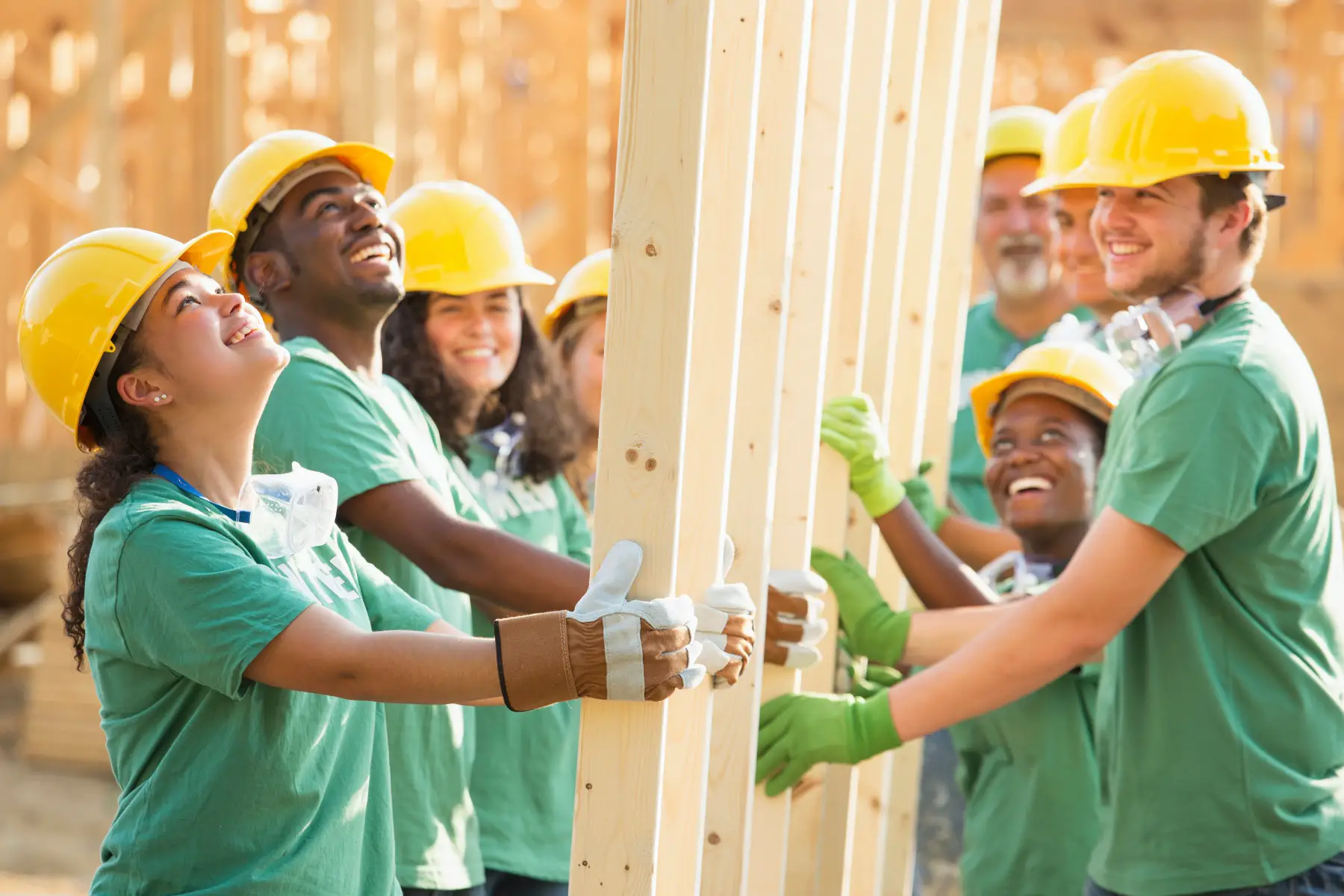 a group of university-age volunteers wearing construction safety helmets and standing in a circle and holding a wooden frame together on a building site