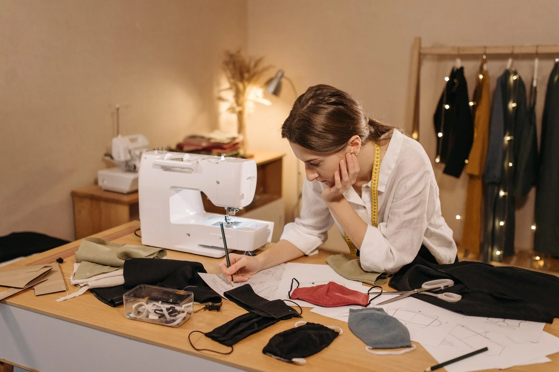 a fashion designer sketches on a piece of paper, next to her is a sewing machine and fabric