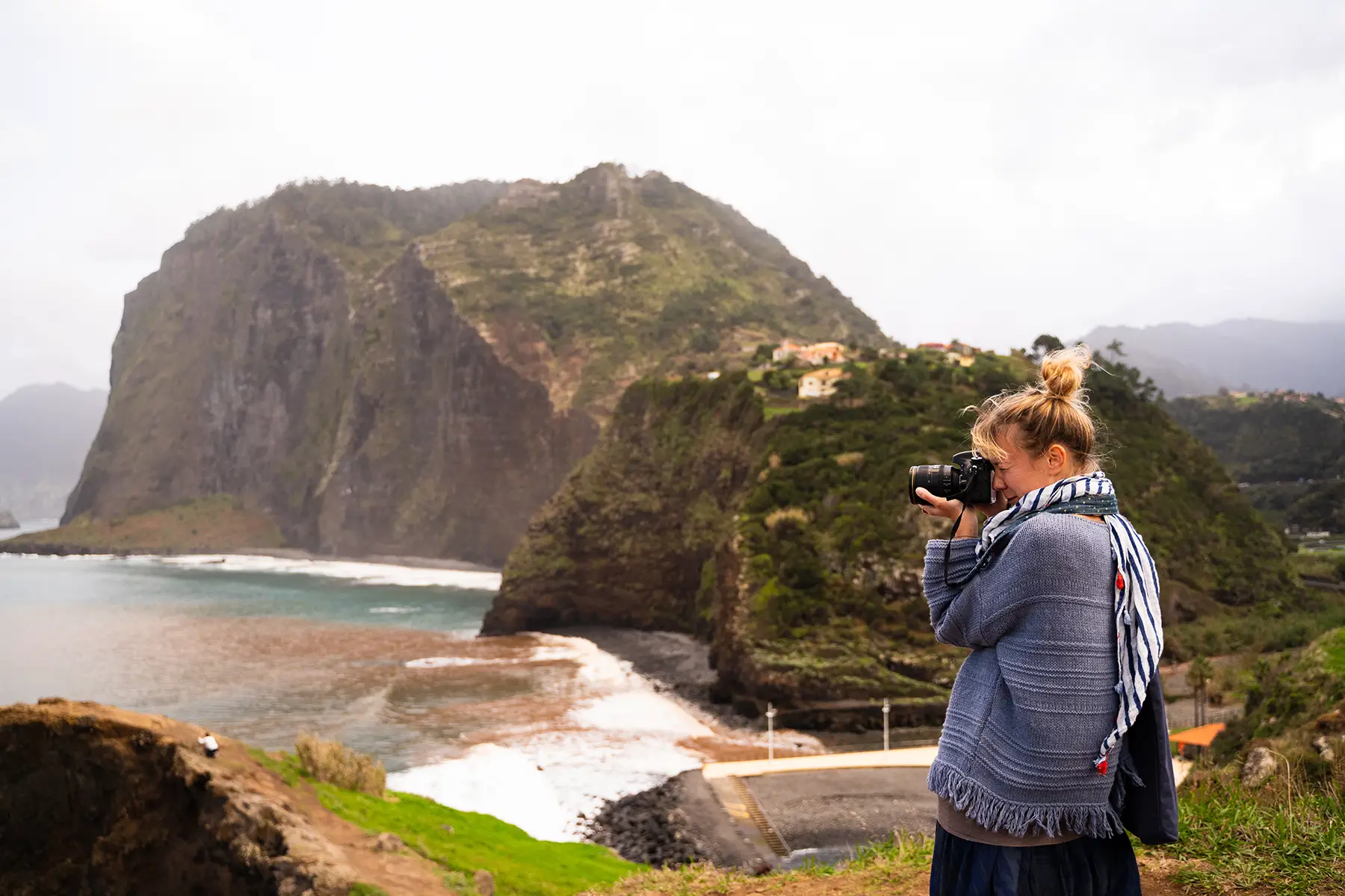 A woman taking a picture on the Portuguese coast, lots of green hills, cliffs, and sea.