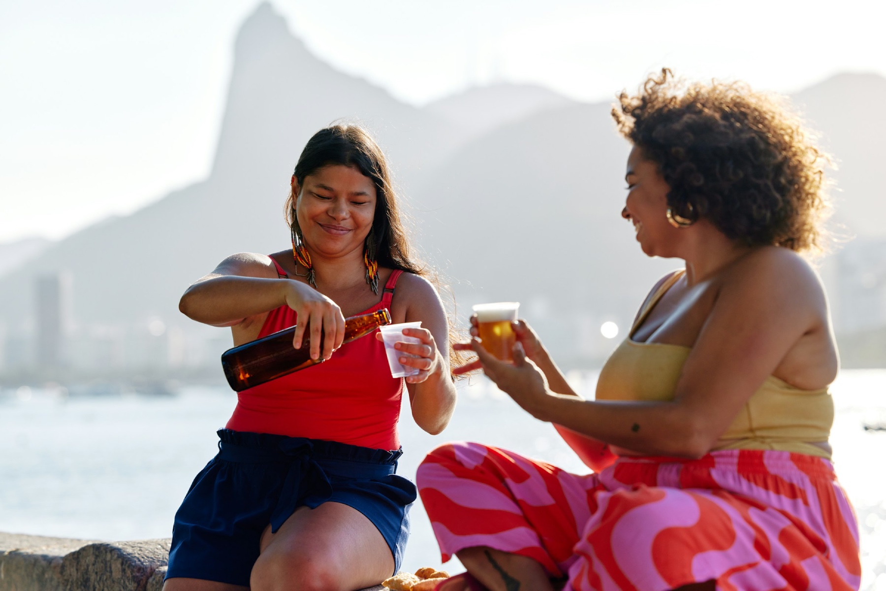 Two woman sit by the sea pouring beer into their glasses, they are relaxed and smiling