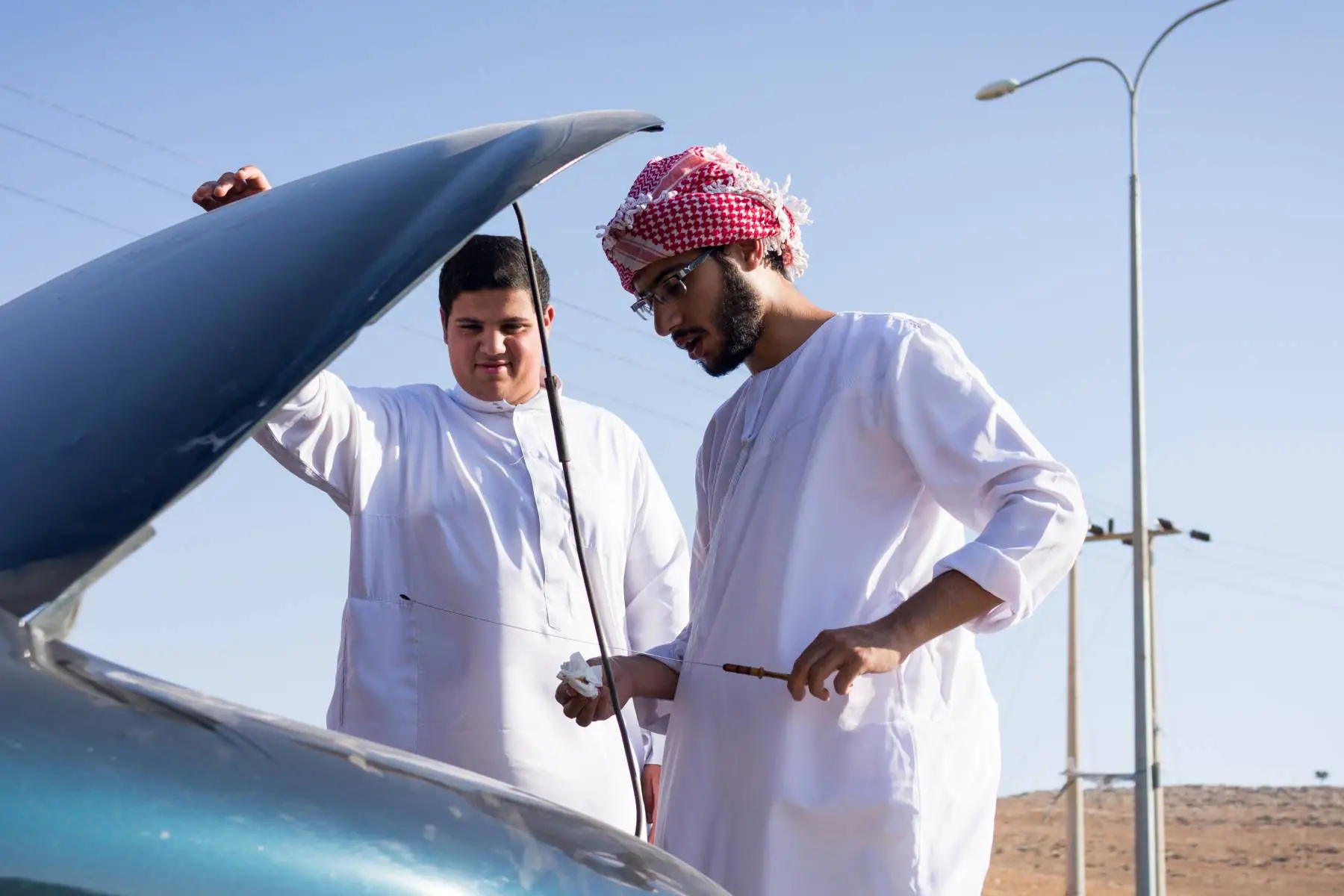 Two young men checking the oil of their car on the side of the road.