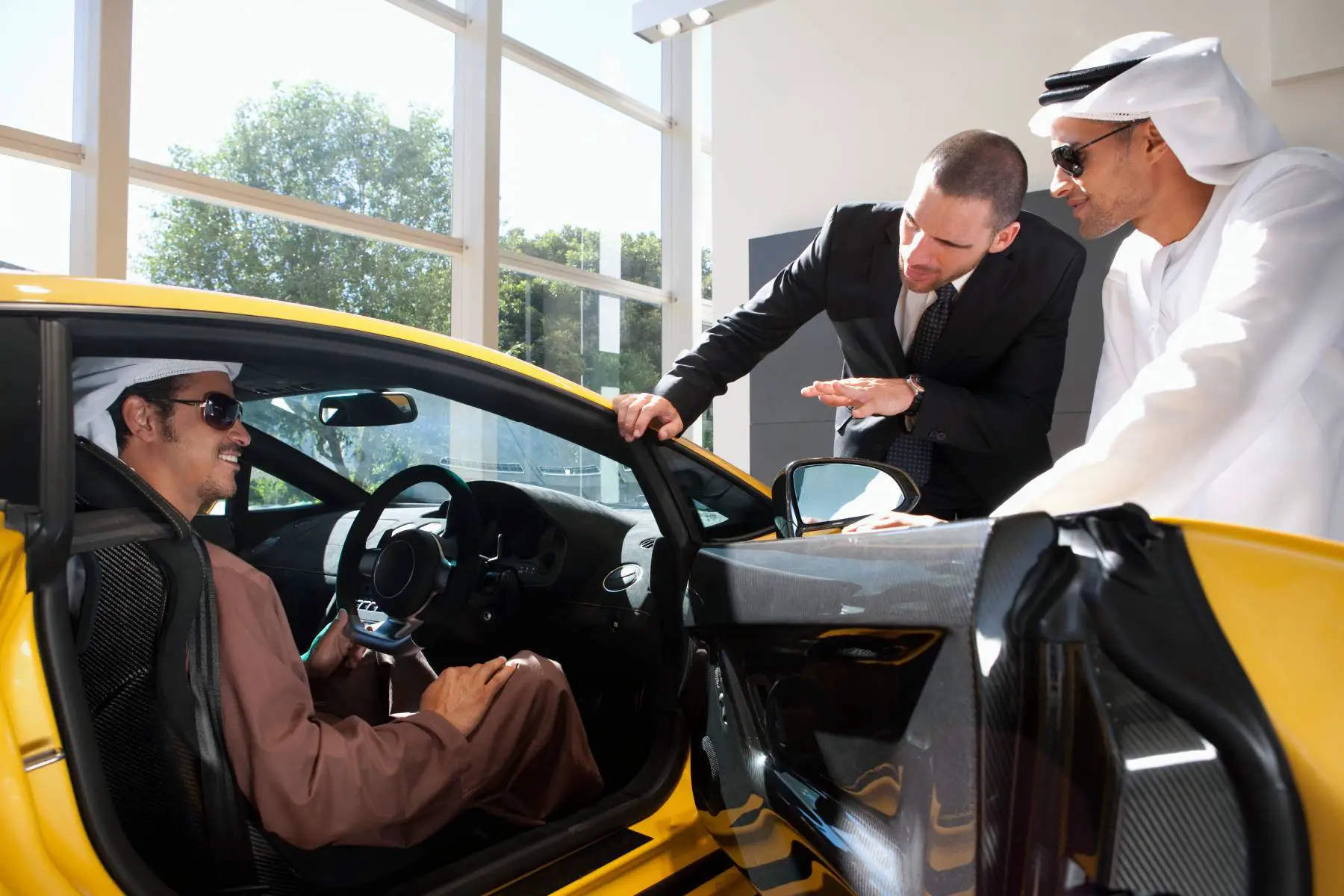 Young man sitting in an expensive car, talking to the dealer. A friend is standing by, holding the car door.