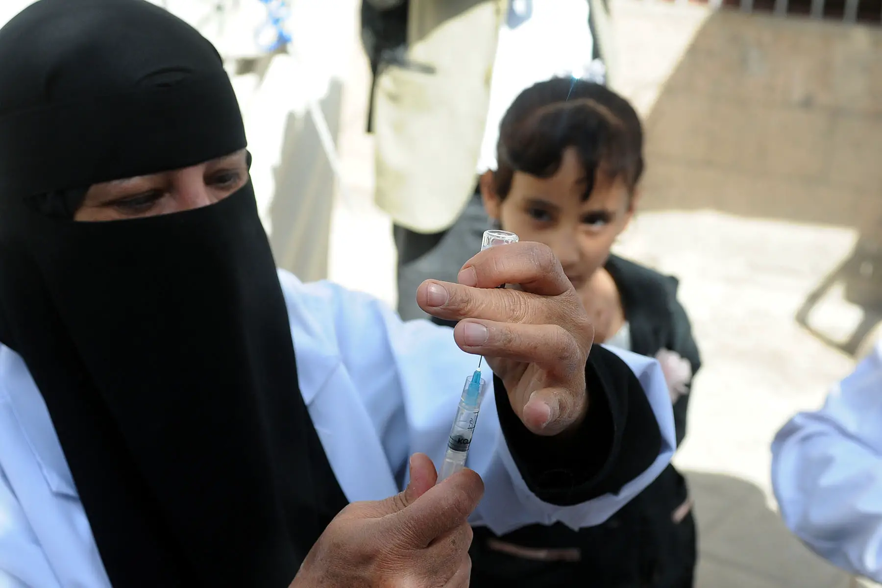 Nurse with a niqab readying a vaccine while a child in the background is looking on curiously.