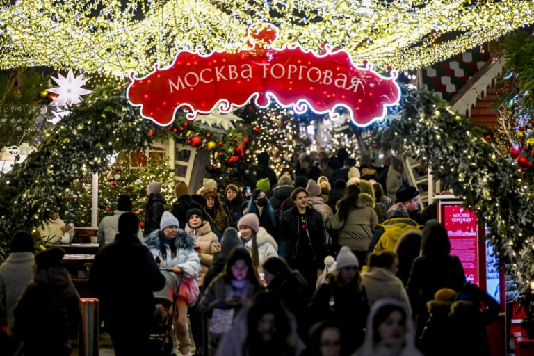 A large crowd of people walk through a Christmas market in Moscow, a banner saying 'Merry Christmas' in Russian hangs overhead