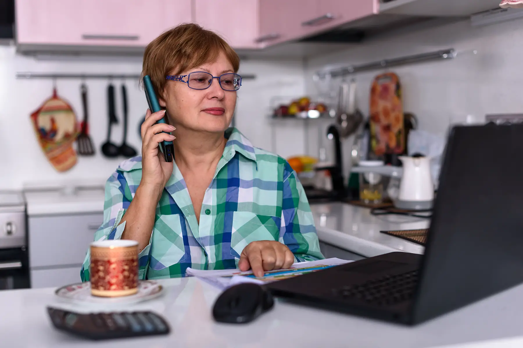 A woman on the phone in her kitchen at a laptop