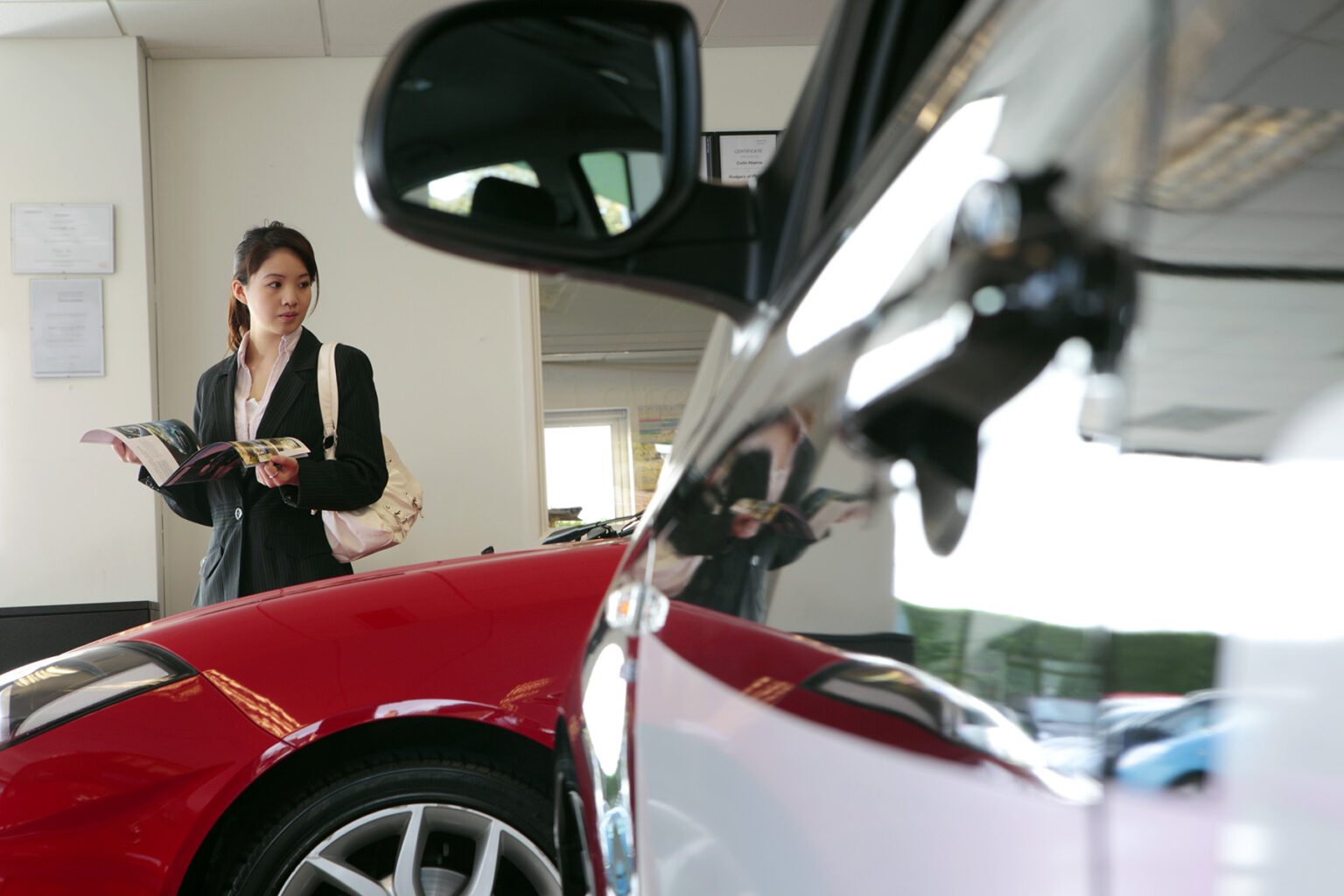 Woman browsing a catalogue as she is looking at a red car inside a dealers showroom.