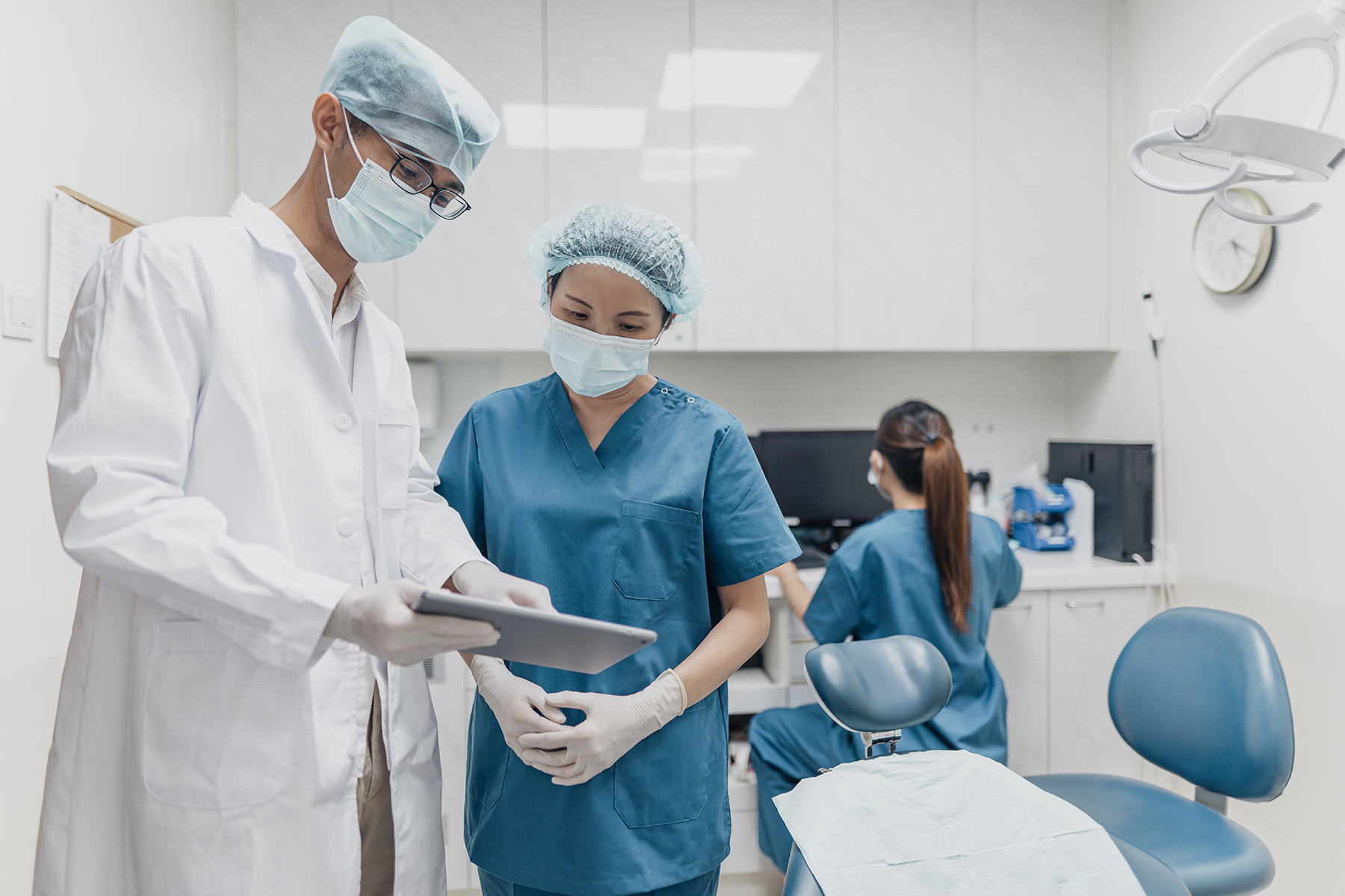 A dentist in a white coat discusses a patient chart with a dental student in a surgery, all wear masks, another dental student or assistant prepares tools for next patient in the background