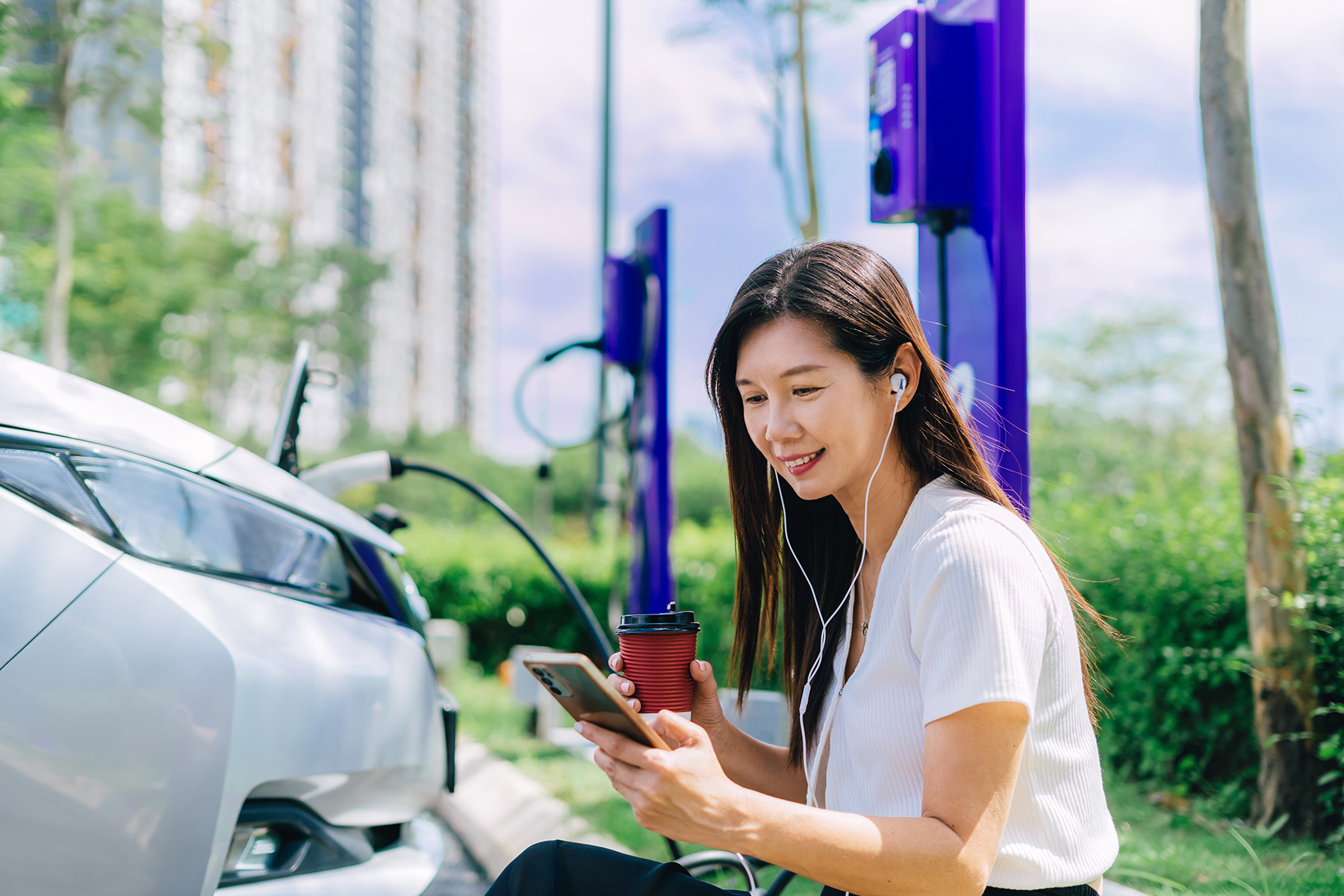 Woman sits next to her EV busy charging, with a coffee and looking at her phone
