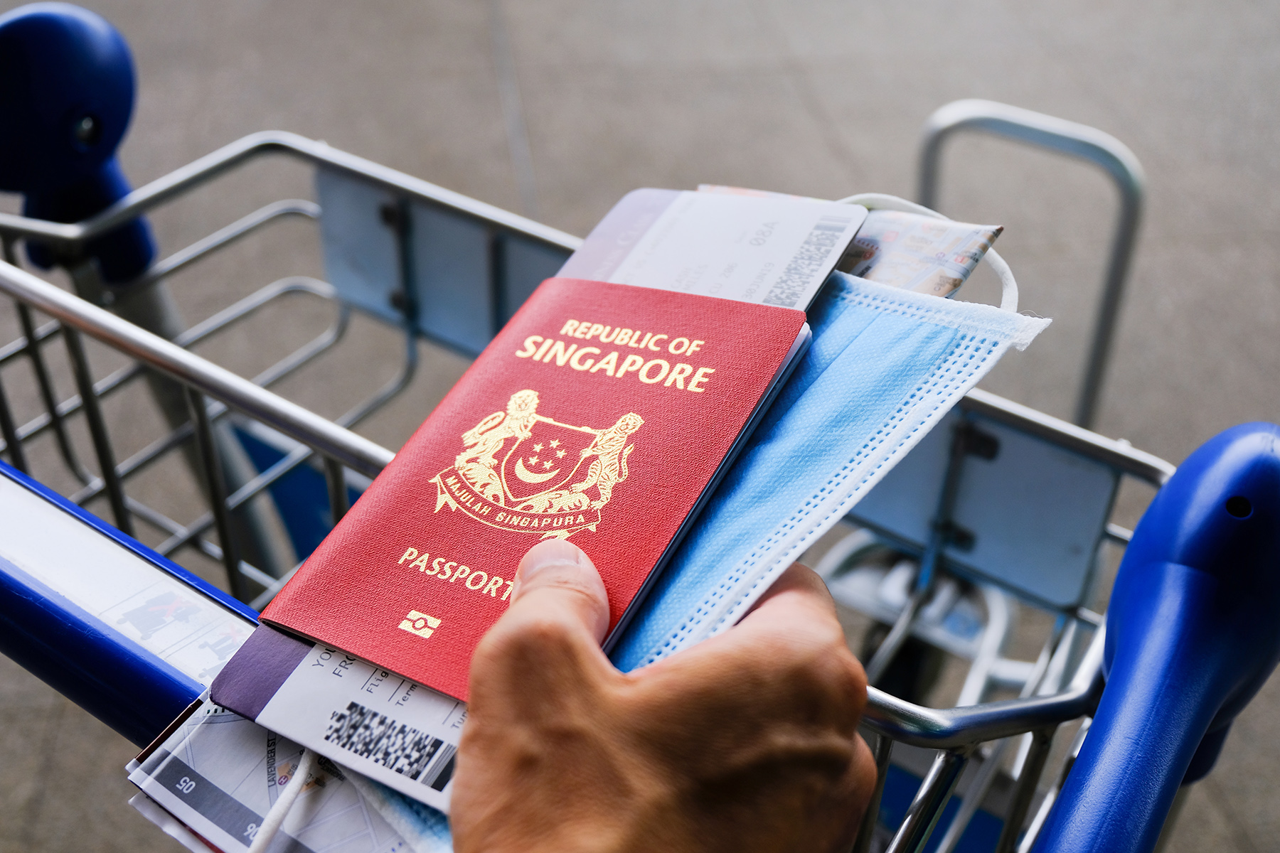 Close up of a hand holding a Singapore passport and boarding pass while pushing a trolley