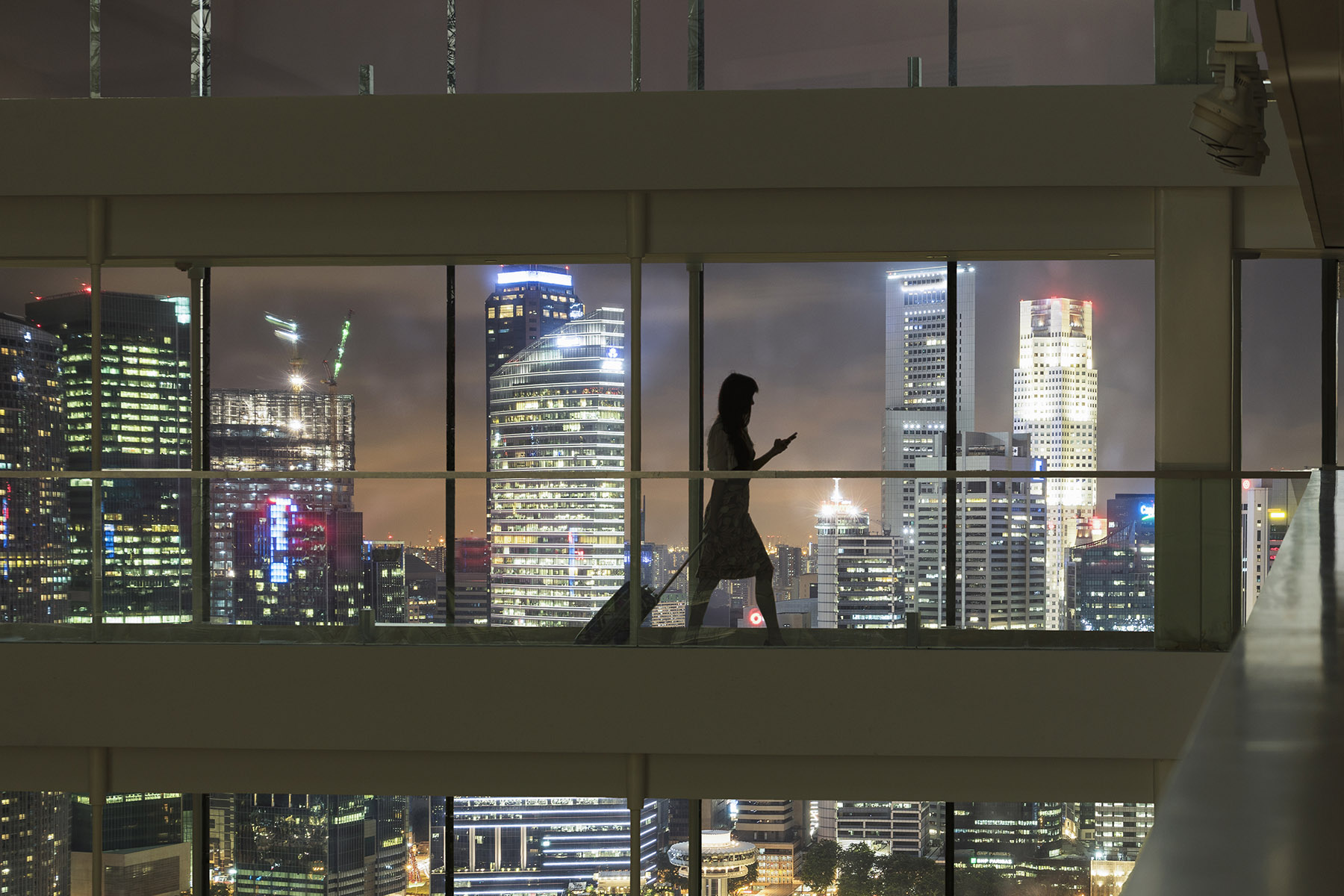 A woman walks alone through a glass paneled sky bridge with a suitcase. She looks at her phone and through the glass the nighttime Singapore skyline is visible
