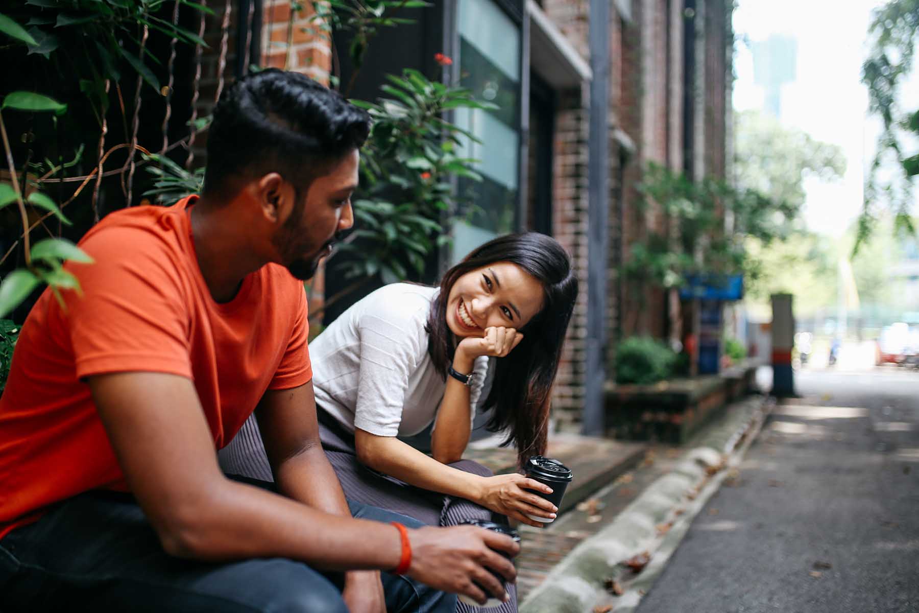Two people sitting outside on a ledge in an alley. Woman is leaning on her hand, smiling like she's in love at the guy next to her. She's obviously flirting.