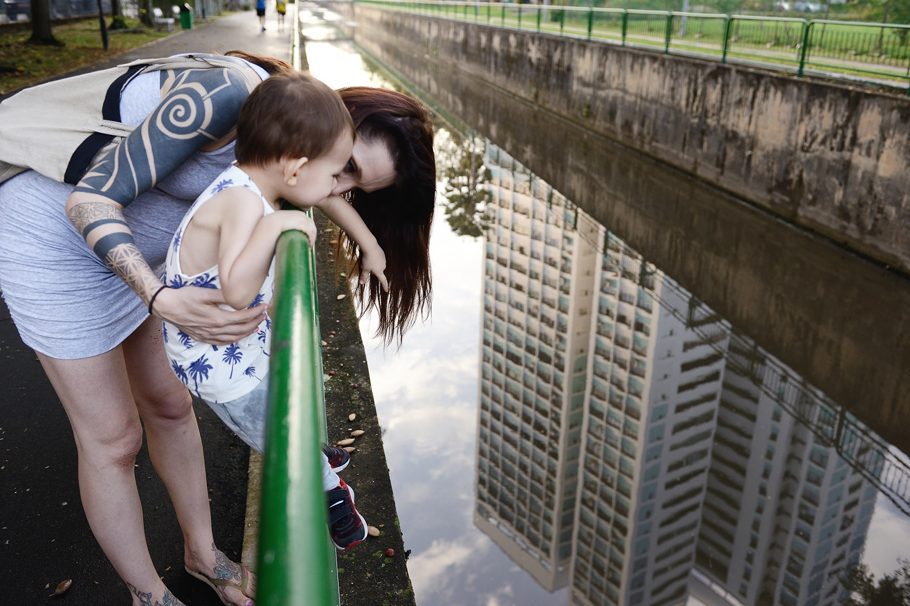 A mom stands with her toddler at a canal, the building's reflection on the water. She has tattoos on her arm