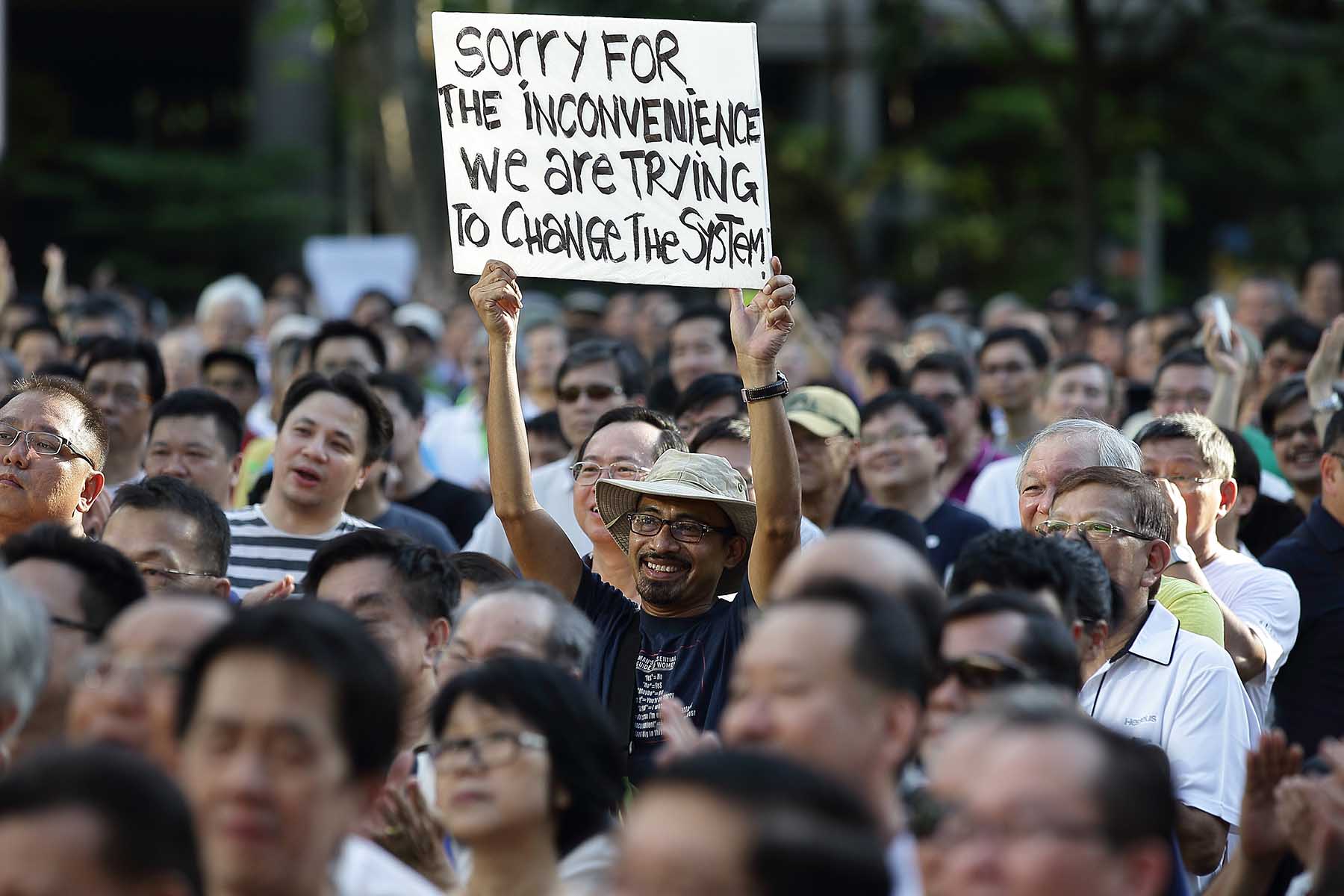 A smiling man holds up a sign saying "sorry for the inconvenience, we're trying to change the system" during a 2014 protest at the Speakers' Corner at Hong Lim Park in Singapore. He is surrounded by a large crowd.