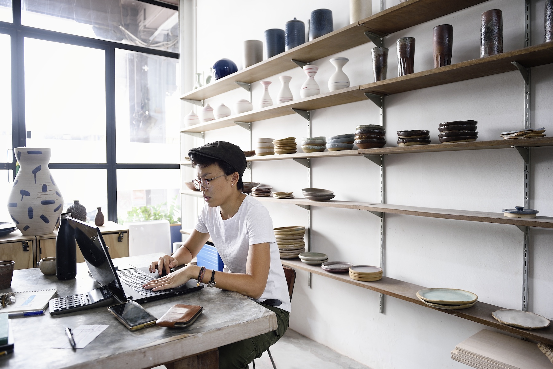 An artist works at their laptop on a desk in the middle of their pottery shop