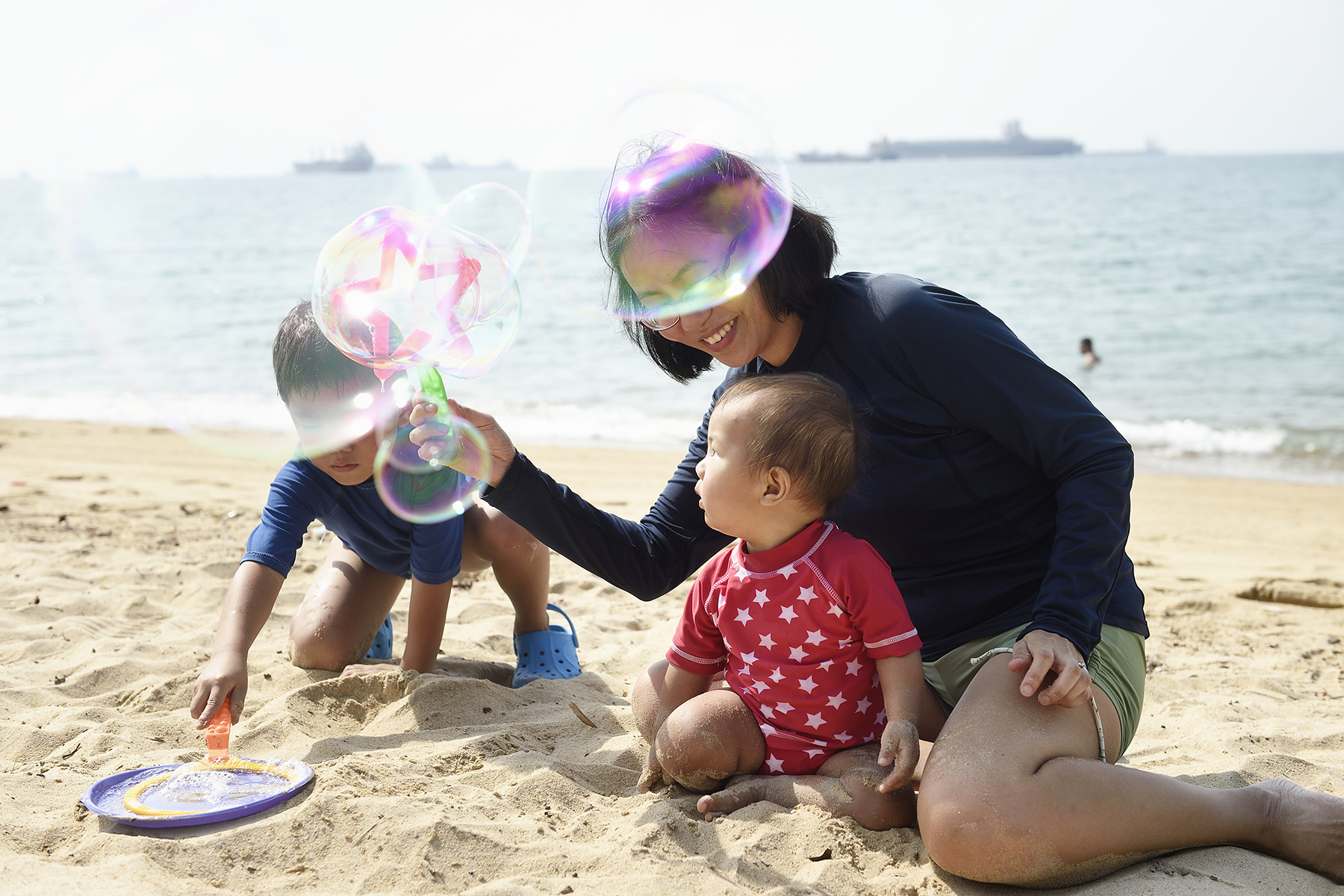 Mother plays with children on beach, blowing bubbles during their first week in Singapore 

