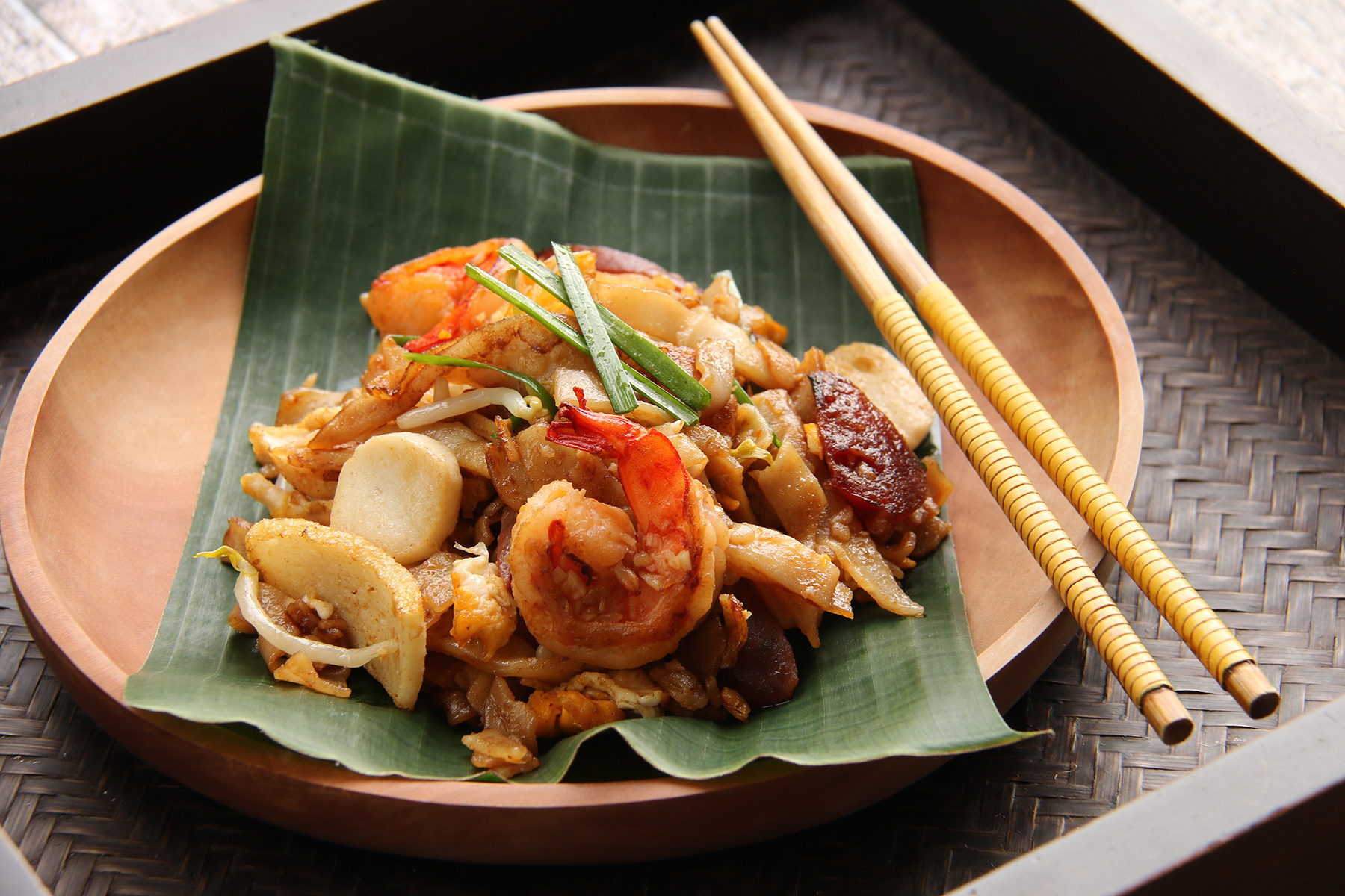 Char Kway Teow, a popular hawker food of stir-fried flat rice noodles from Singapore and Malaysia. It is served on a wooden plate that has been lined with banana leaf and chop sticks