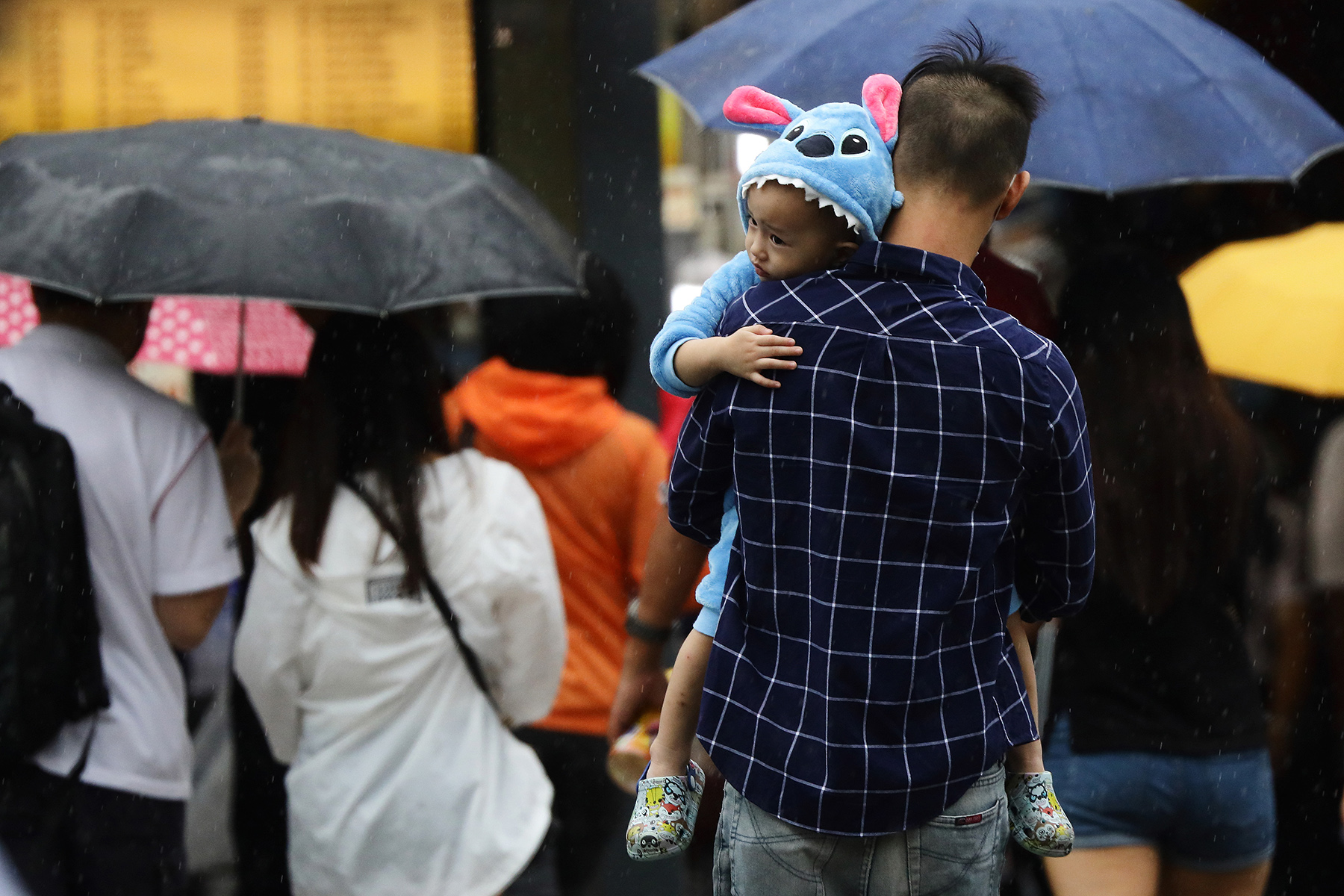 From behind, a dad carrying his child who is wearing a blue onesie - it is raining, other people have umbrellas, she can join him in Singapore on a Dependent Pass

