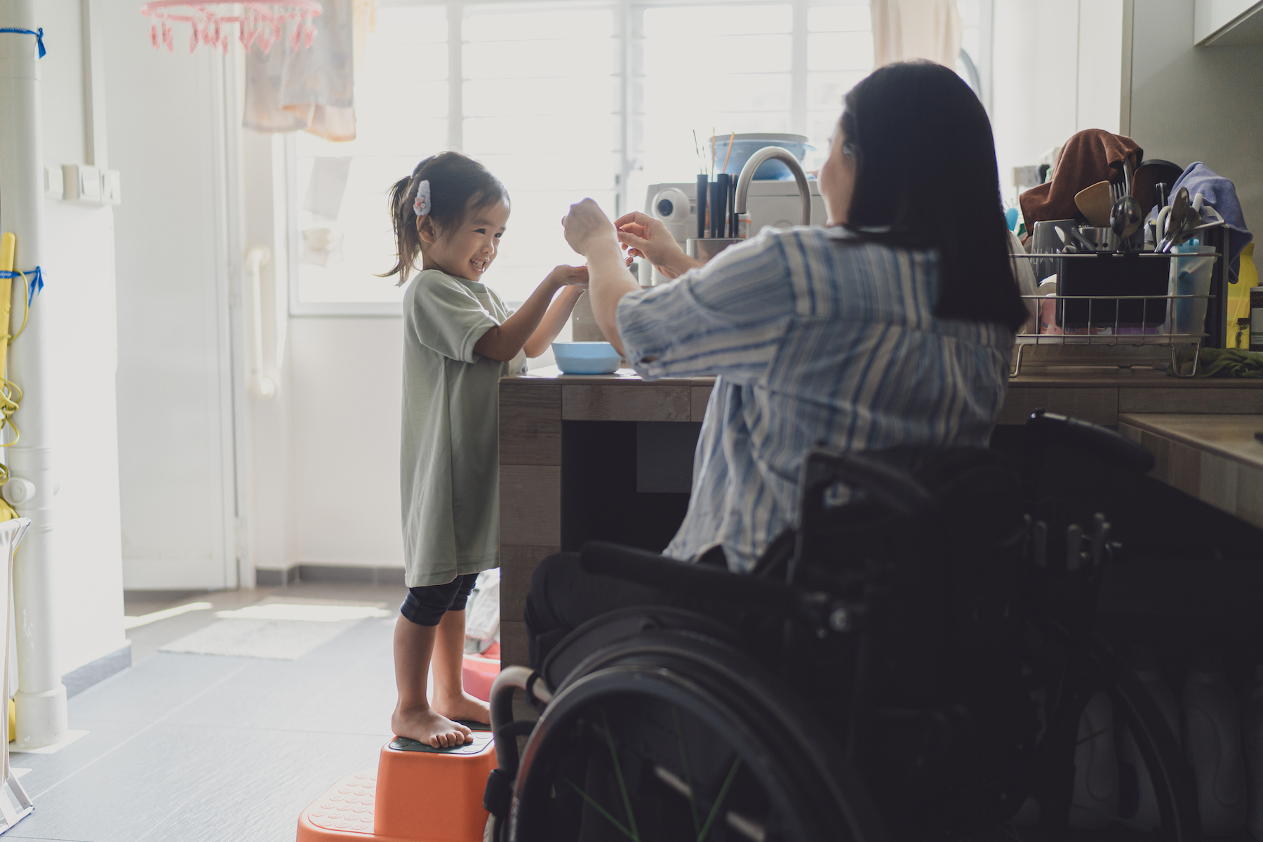A mother in a wheelchair plays with her young daughter at the kitchen sink
