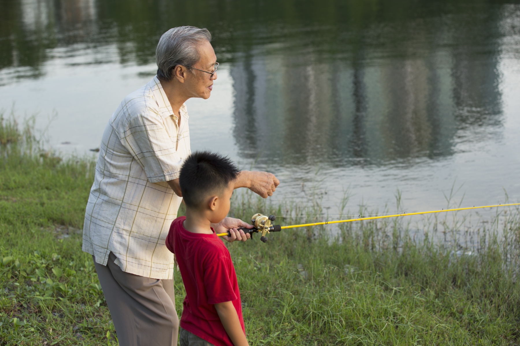 A grandfather teaches his grandson how to fish at the edge of a pond