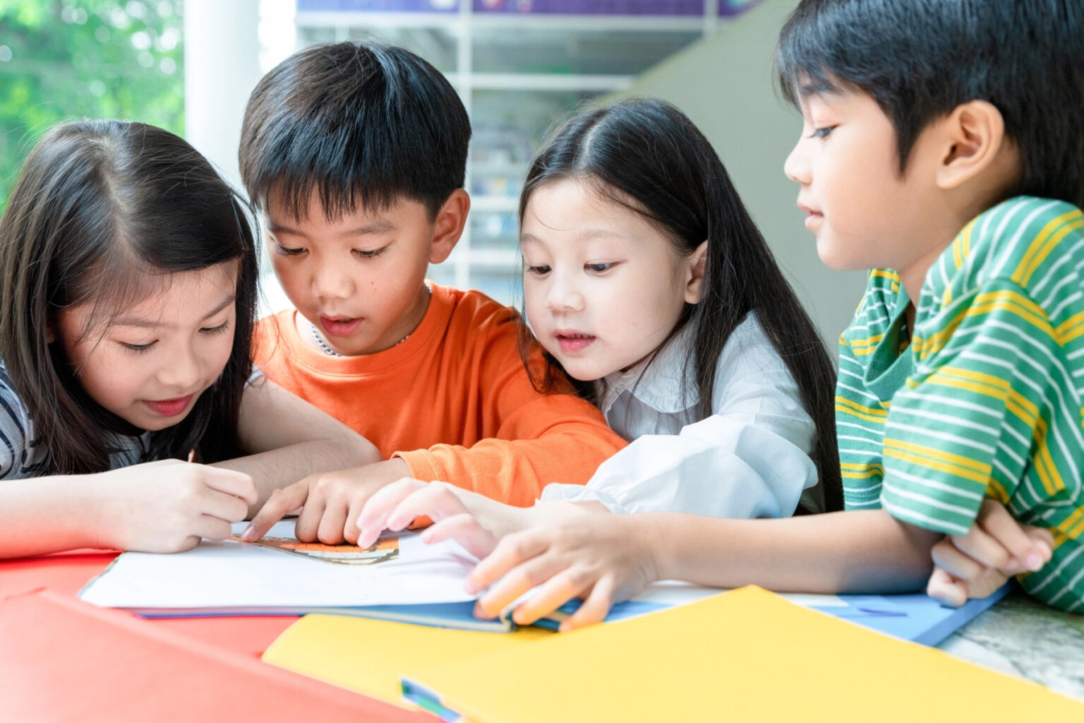 Four Asian children standing around a colorful table look down and reach for a paper