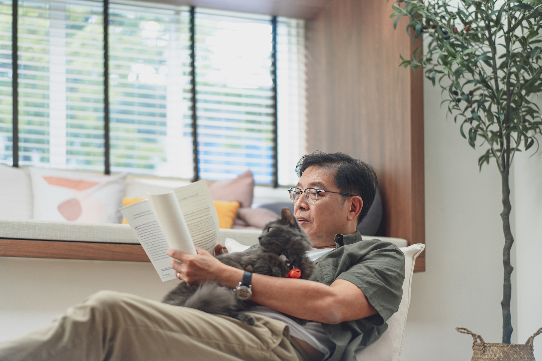 An older man sits in a chair reading a book with his cat on his lap