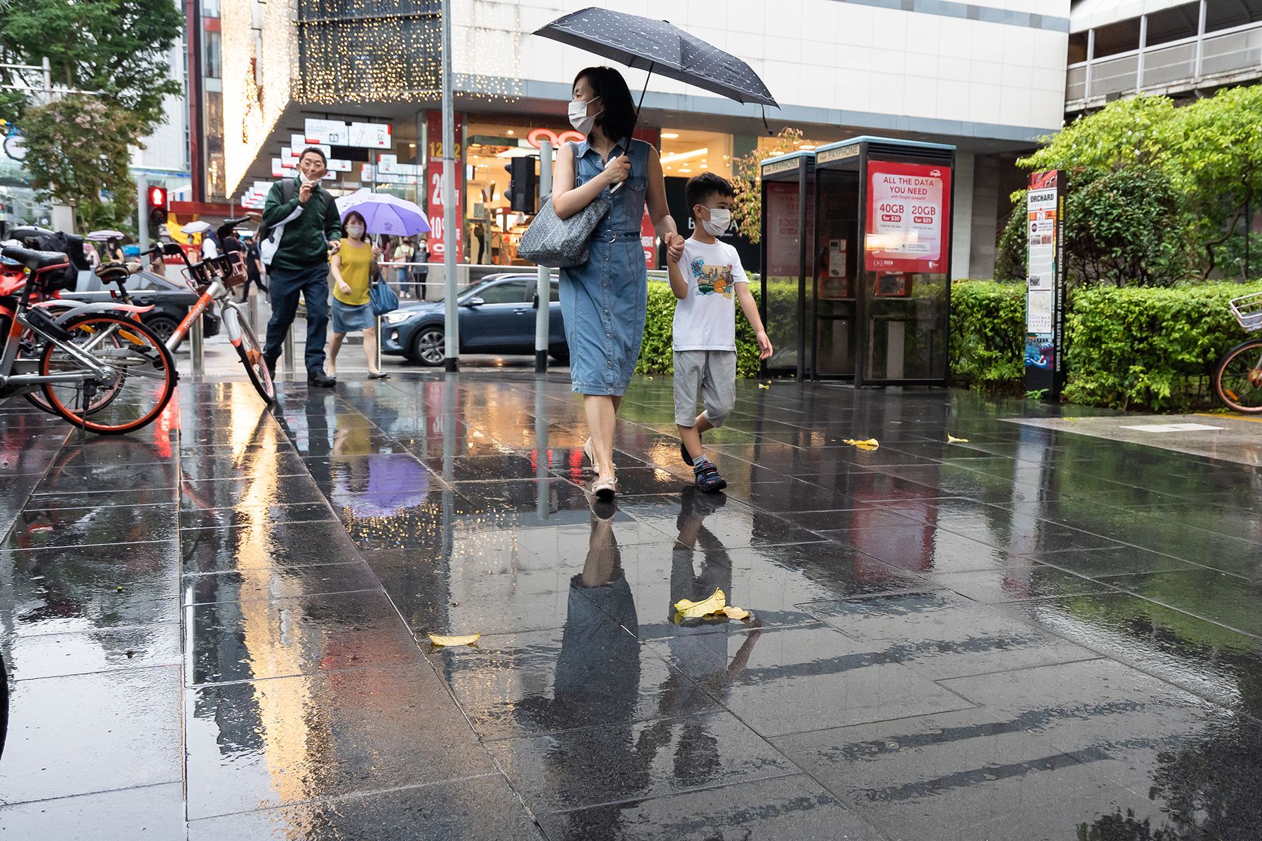 Mom holdd her child's hand while they cross the street, she also hold an umbrella and they are wearing facemasks

