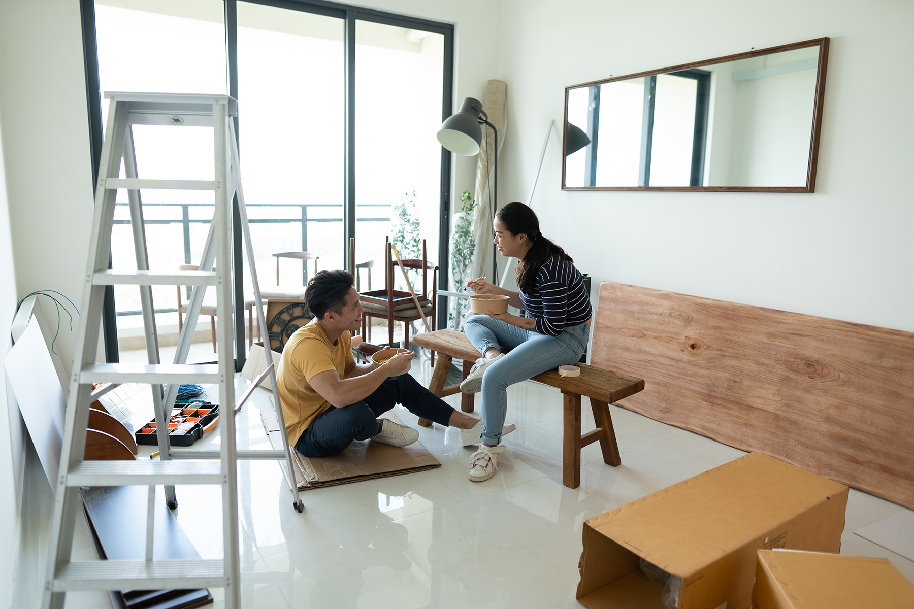 Couple sitting in their empty new home, boxes and ladder standing around, eating lunch

