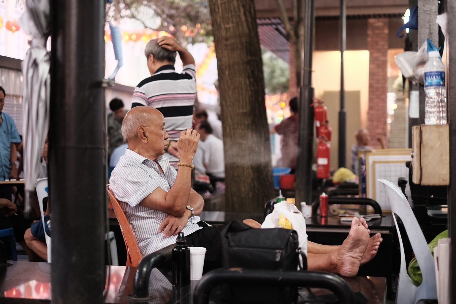 An older man relaxes outside, sitting on a bench or at a café - bare feet on a chair, enjoying a smoke