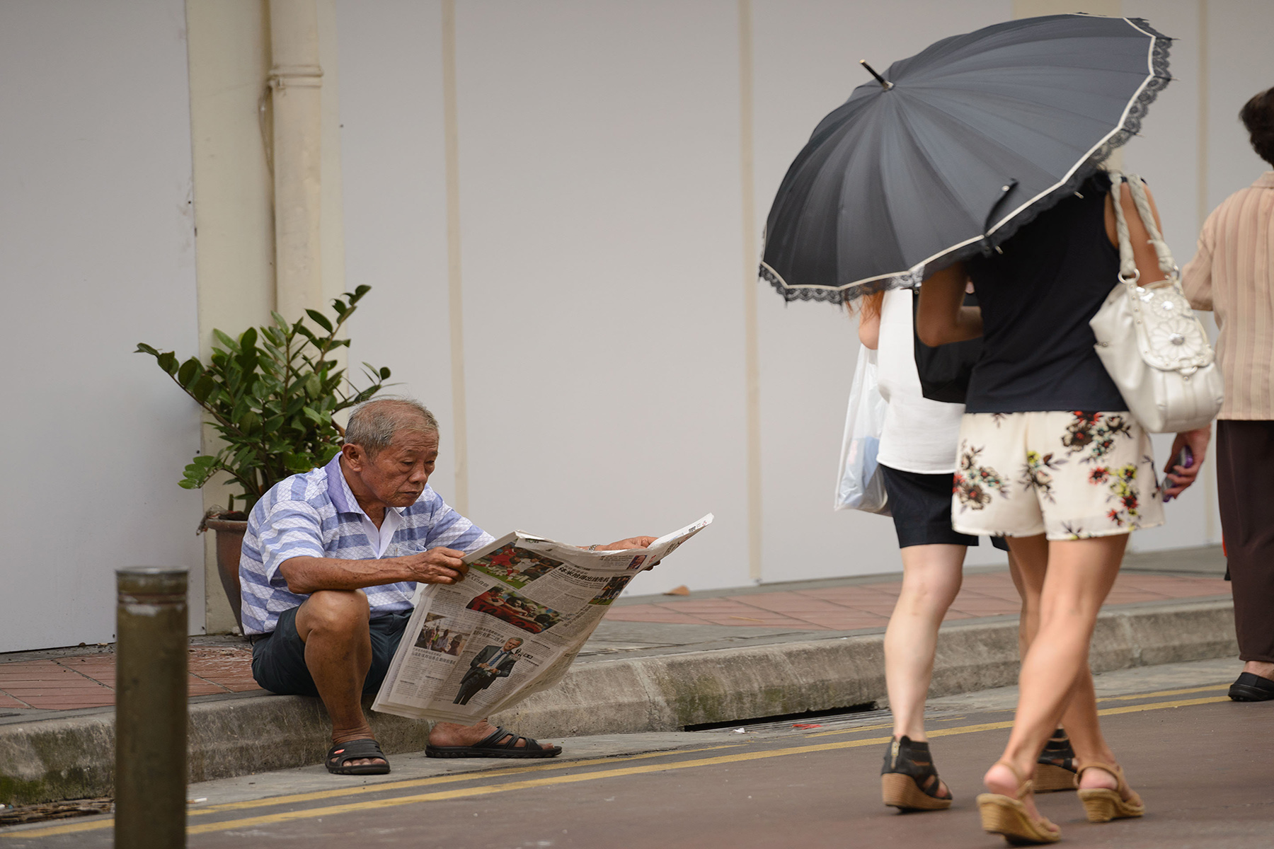 Senior man sitting on the sidewalk, relaxing and reading a newspaper, two people walk past him holding an umbrella

