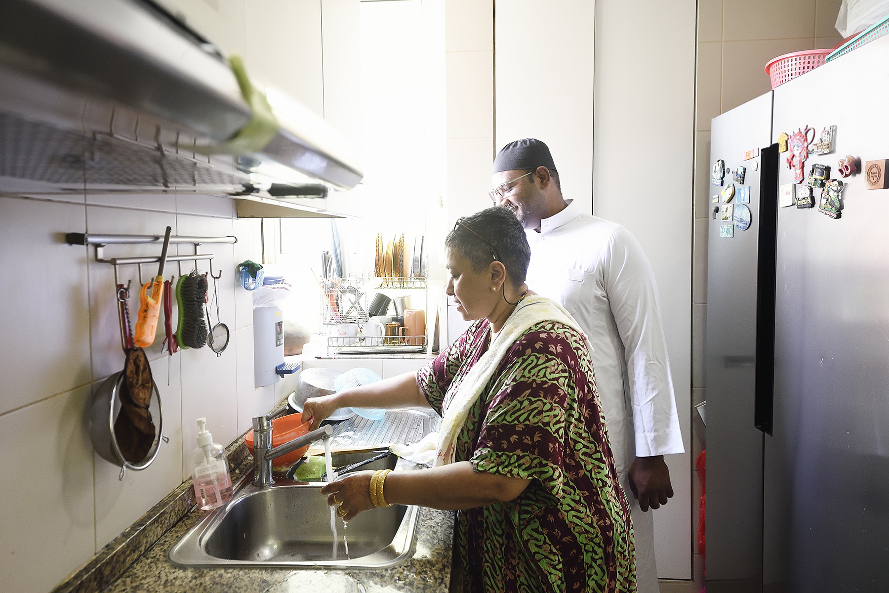 Mother is washing the dishes in a kitchen, while her grown-up son is laughing and doing nothing.