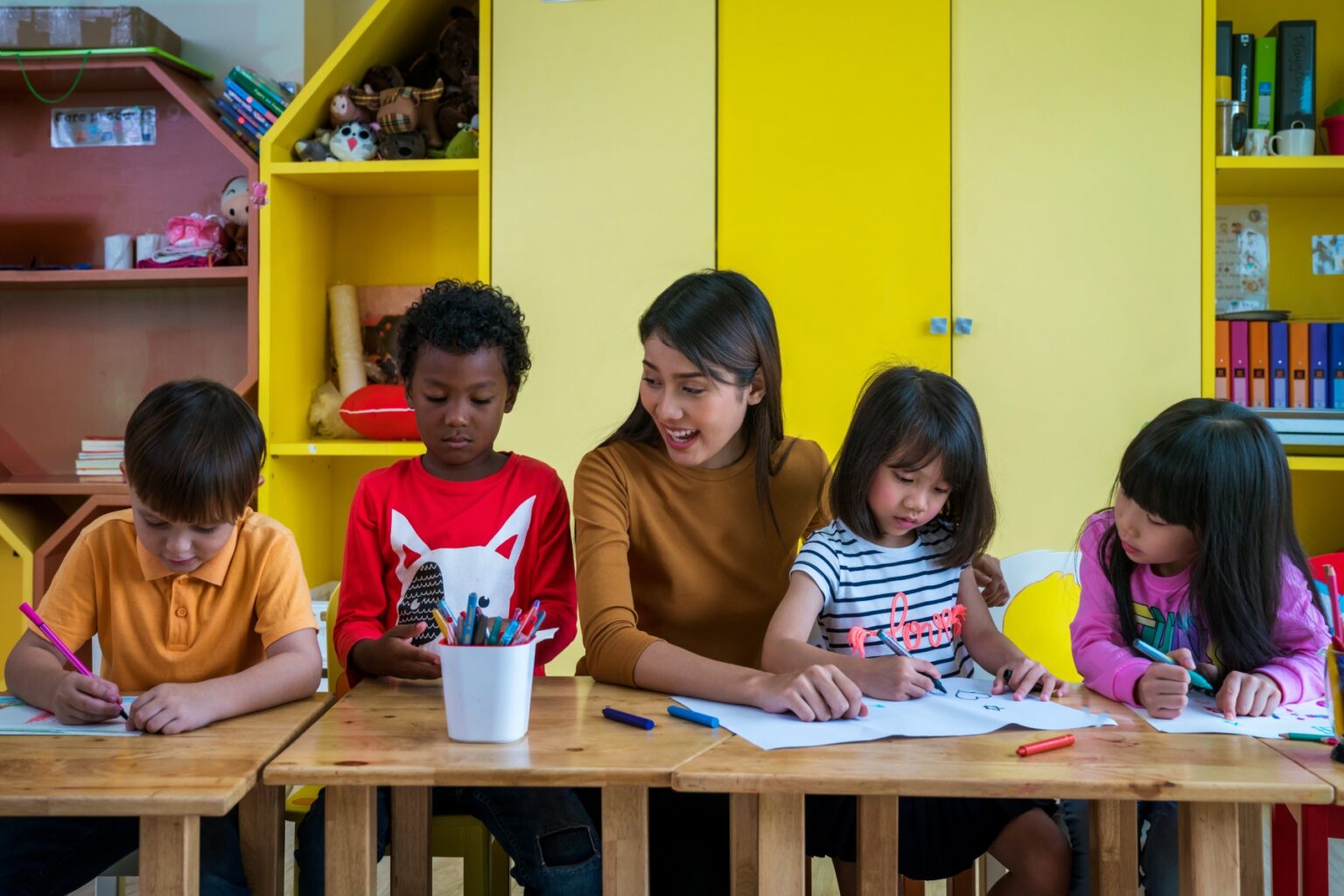A teacher helps four young students of various ethnicities to color on paper in a brightly lit classroom