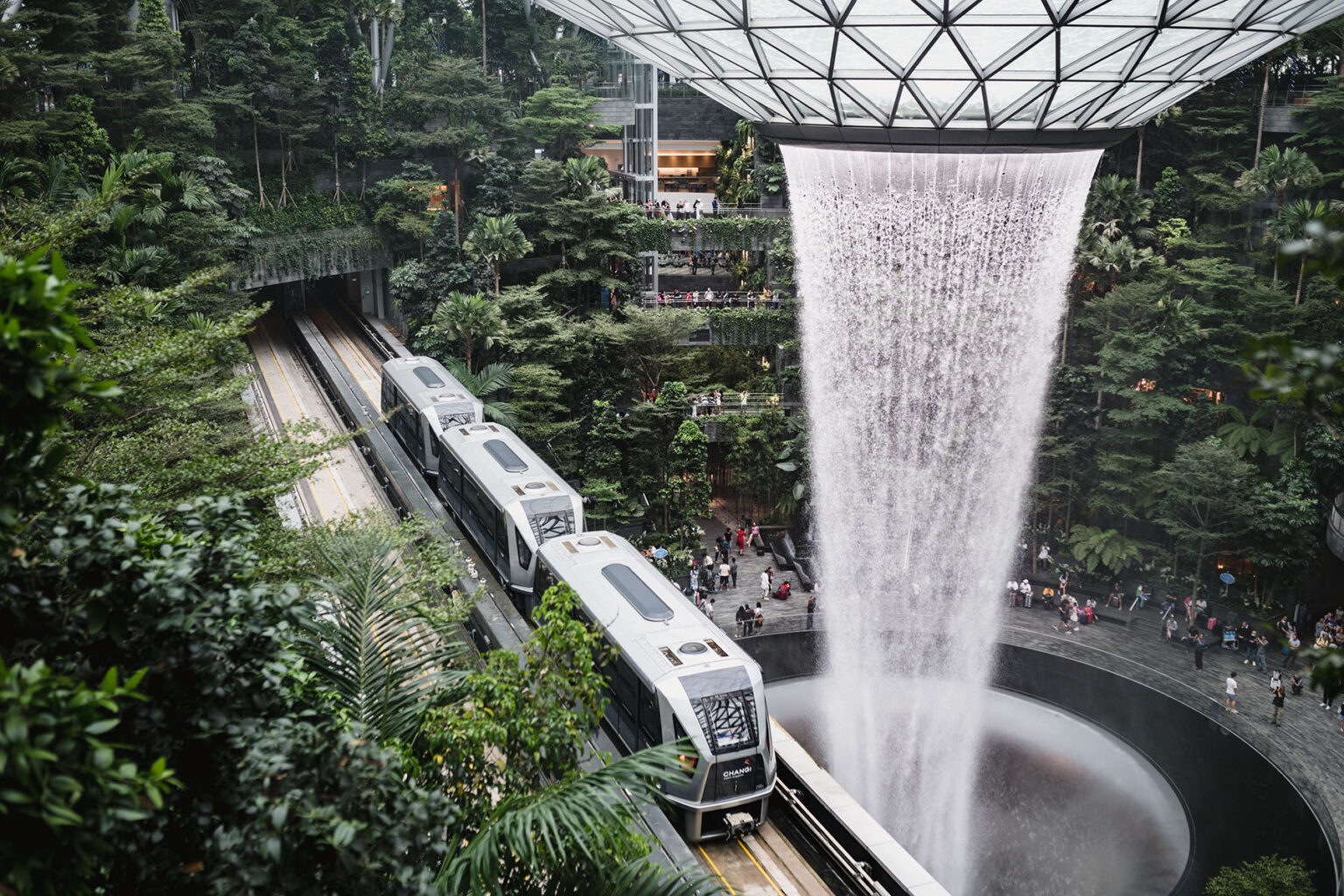 The HSBC Rain Vortex in Jewel Changi Airport is a massive waterfall pouring own. It is surrounded by green nature and a metro is driving by.