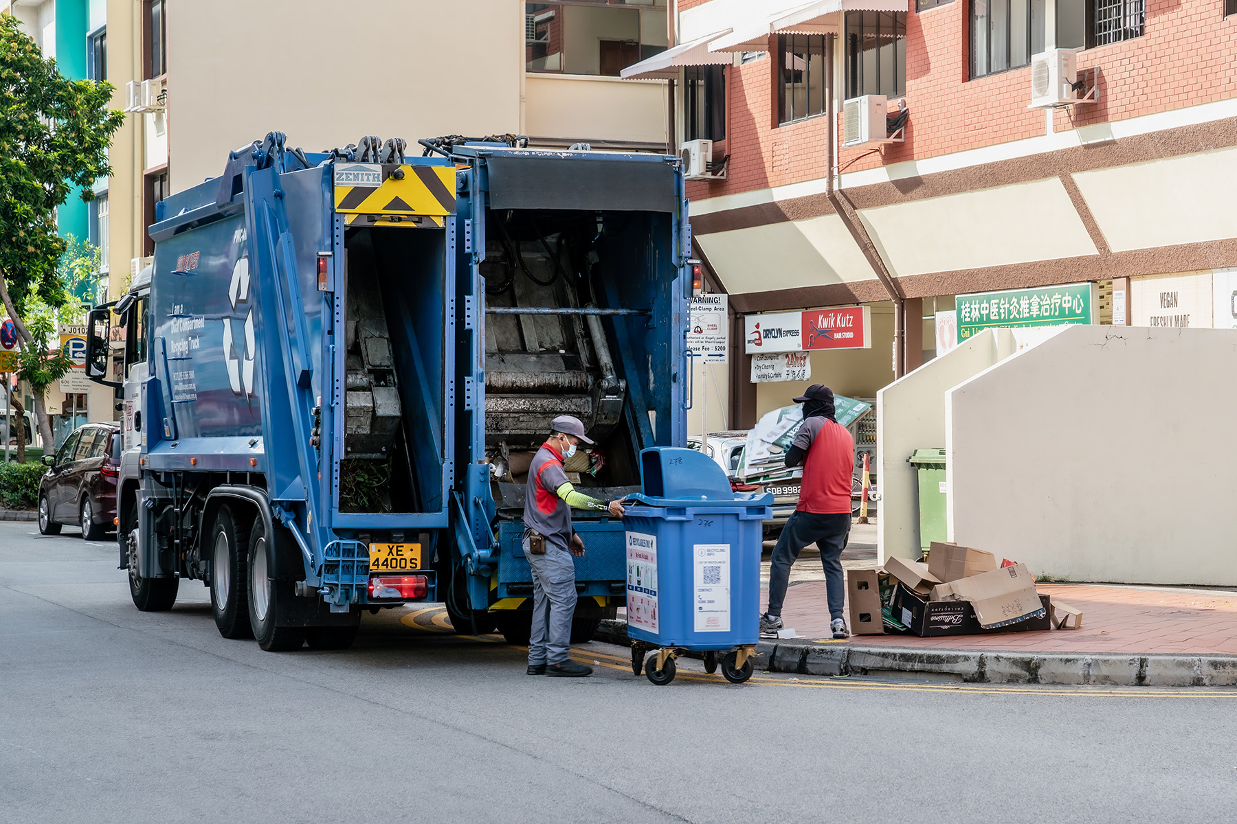 Two garbage collectors picking up general trash with their blue waste management truck in Singapore.