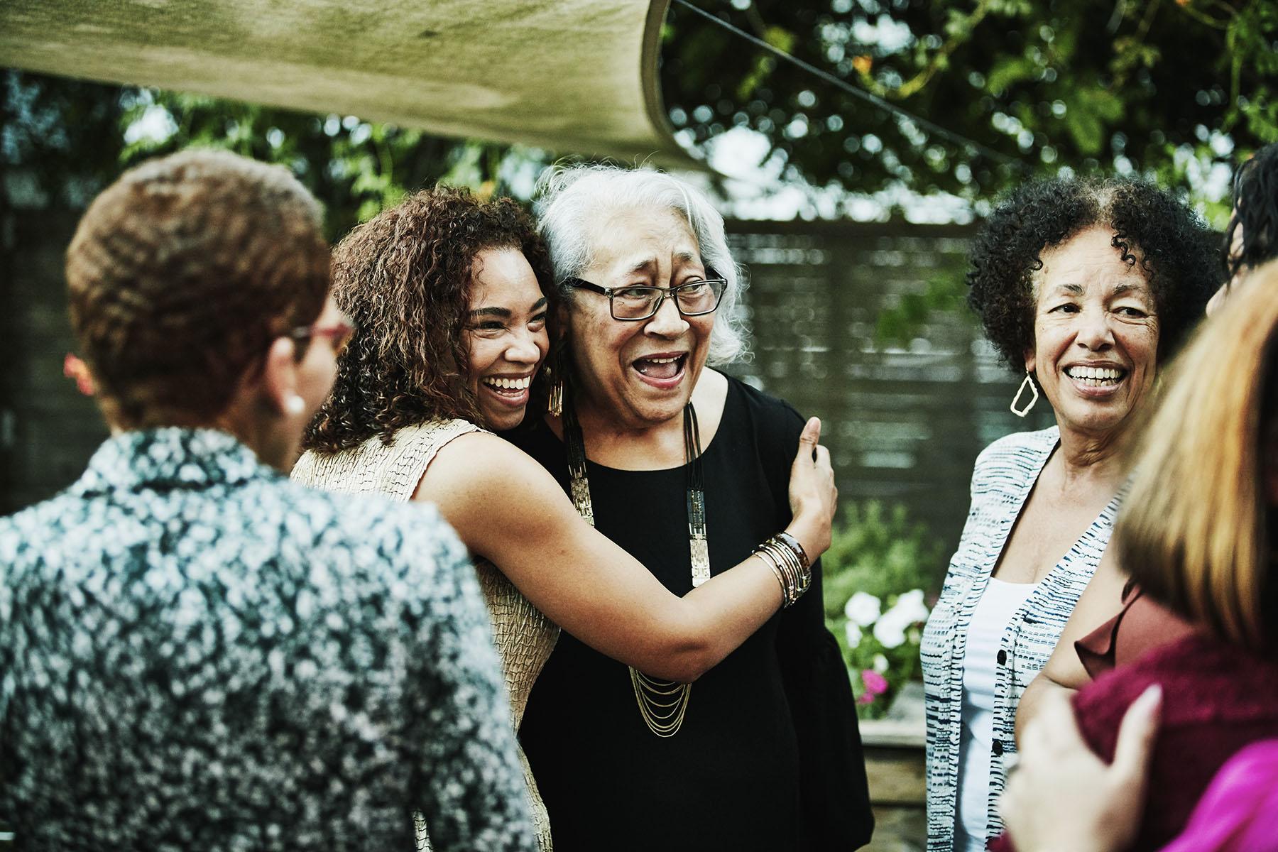 Woman embracing elderly (grand)mother while standing outside in the garden. There is a small circle of people, everyone is smiling and laughing.