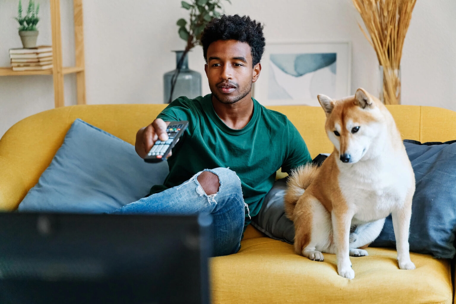 A man sits on the couch beside his dog, turning on the television via remote