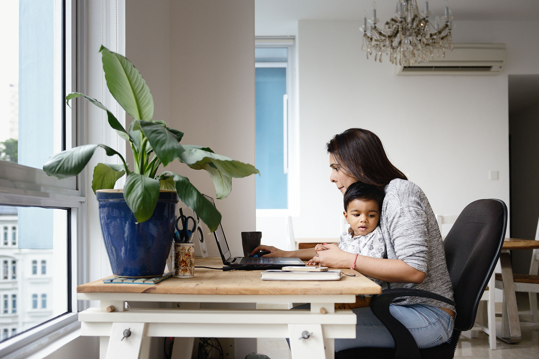 Woman sitting at a desk behind a laptop, while her son sits on her lap. They're both in a modern looking apartment.