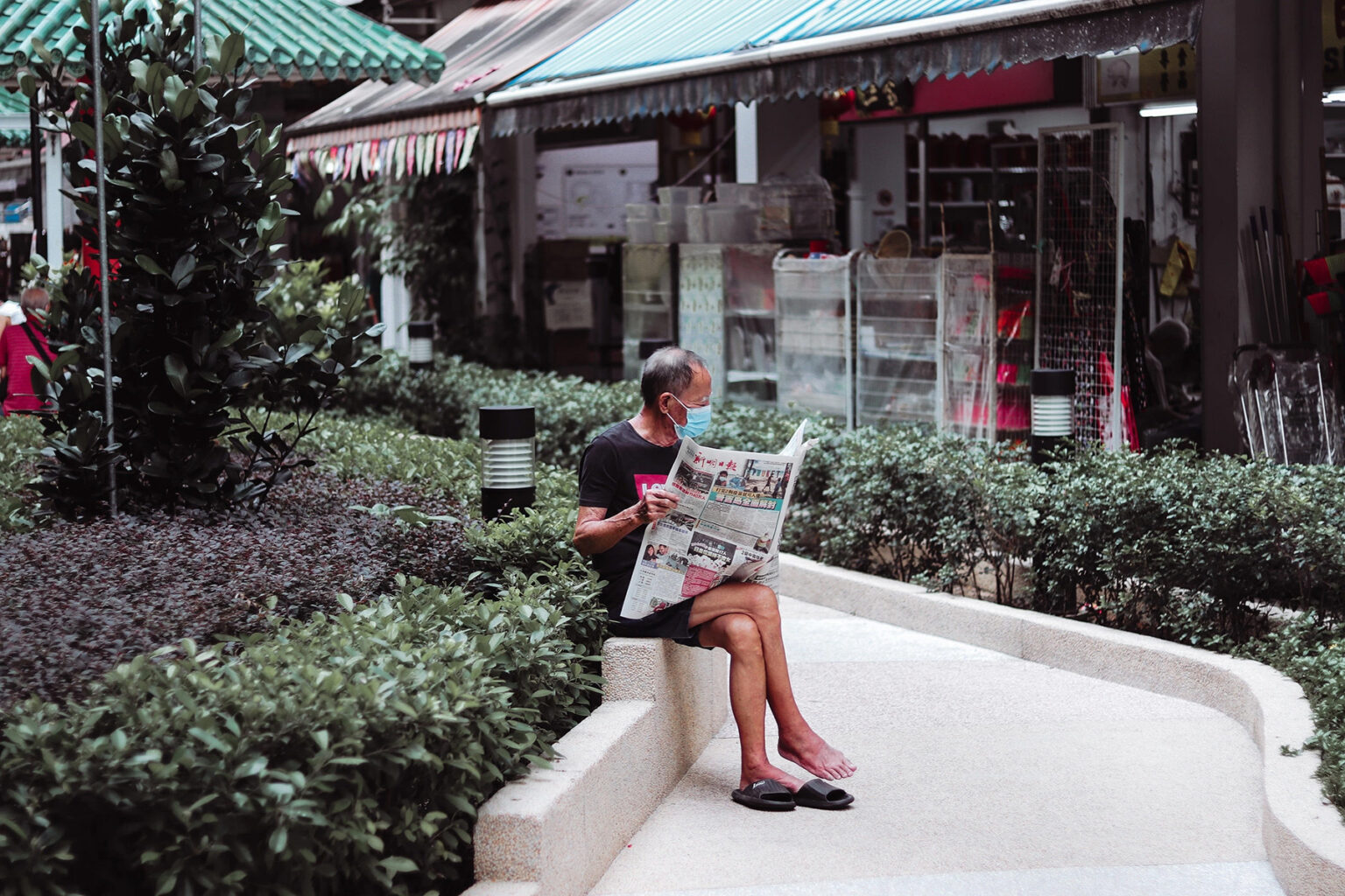 A man reading a newspaper sitting on a wall outside a shop