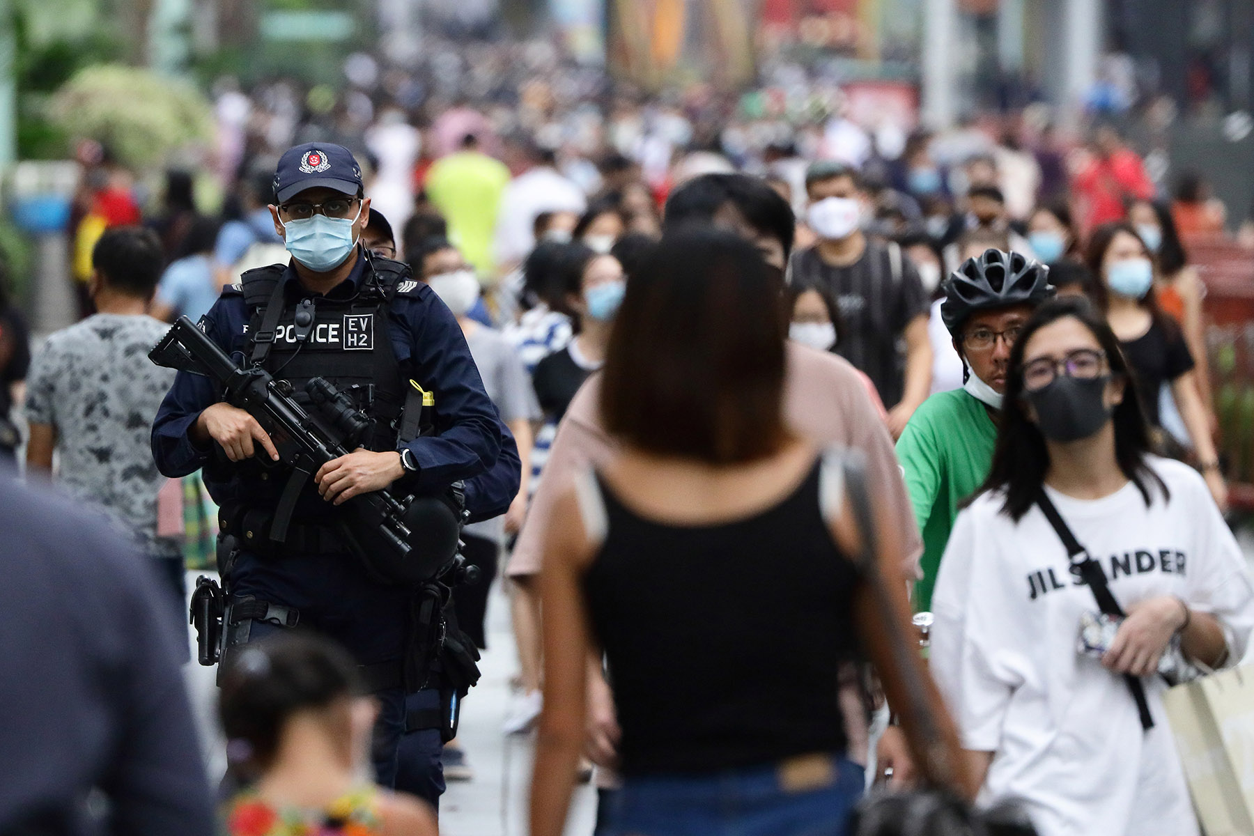 Police officer with glasses and a face mask is holding a huge gun while patrolling in a busy crowd.