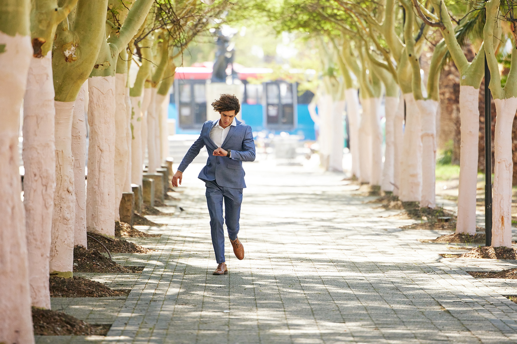 Young man in formal wear running down a lane with trees while looking at his watch. He's late for something.
